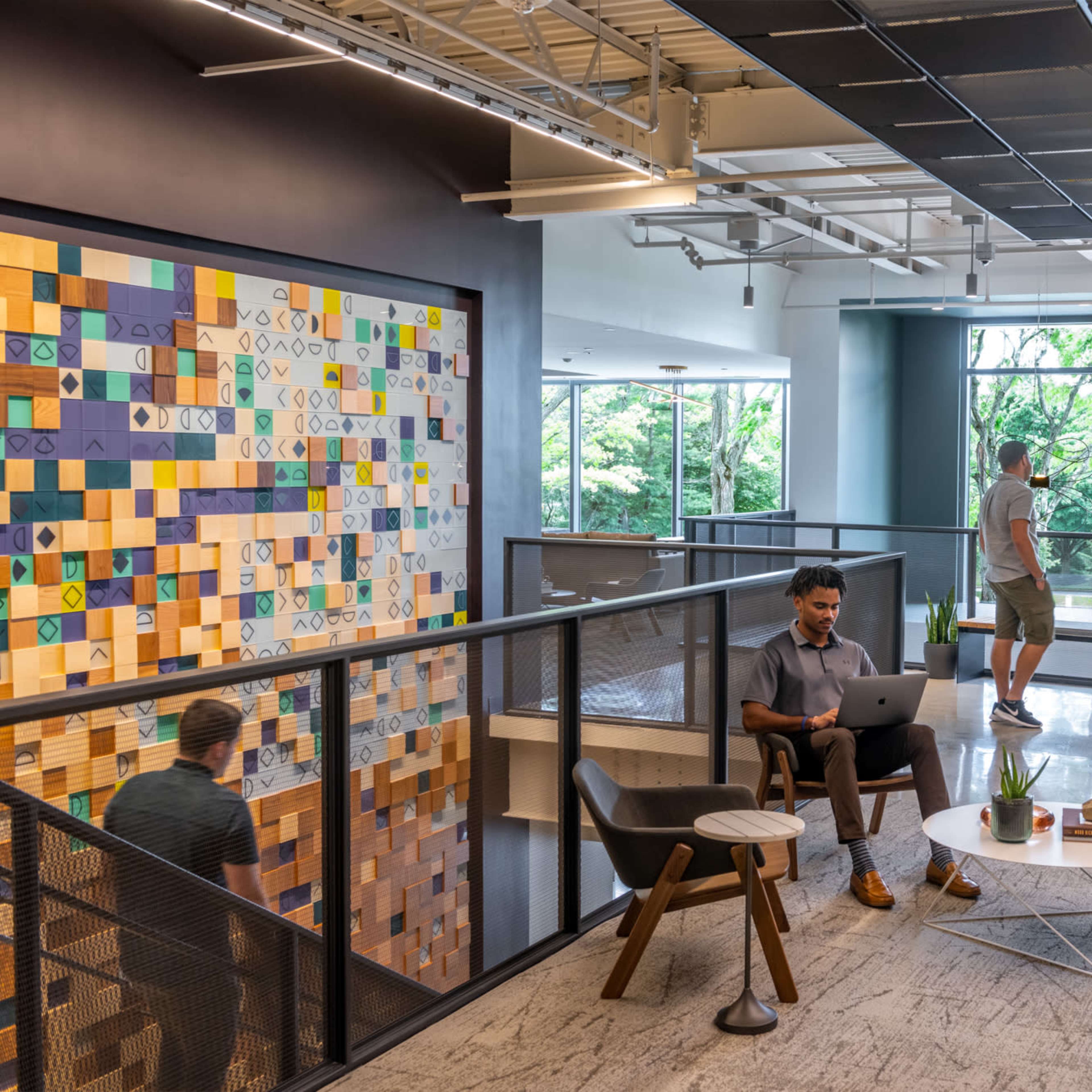 A modern office space with diverse individuals engaged in various activities, including a person working on a laptop and others interacting near large windows that provide natural light and view of greenery outside.