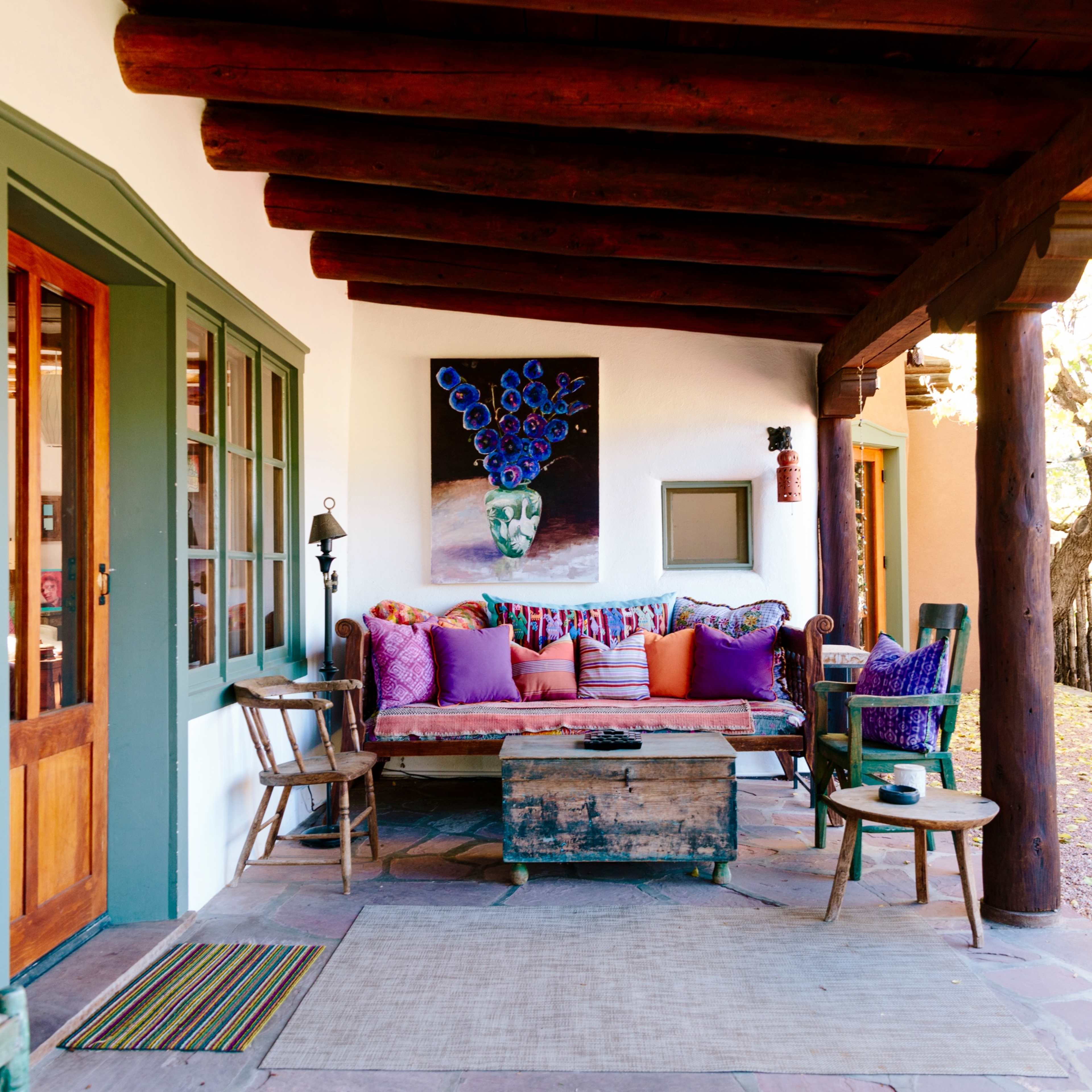 A cozy outdoor seating area with a wooden bench adorned with colorful cushions, surrounded by rustic chairs and a table under a wooden porch.