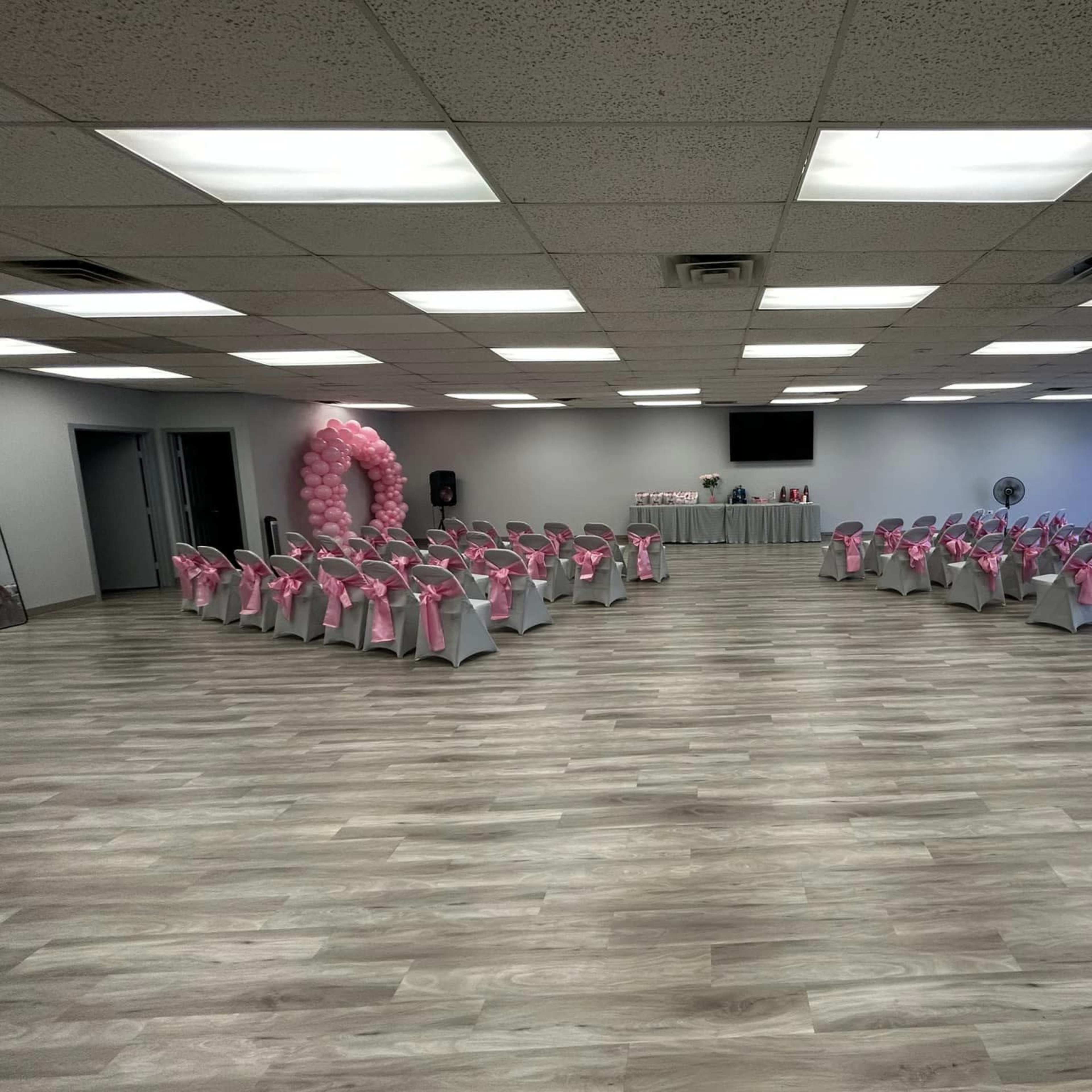 A spacious event room arranged with chairs covered in white cloth and pink sashes, set up in two rows facing a central area decorated with balloons and a table.