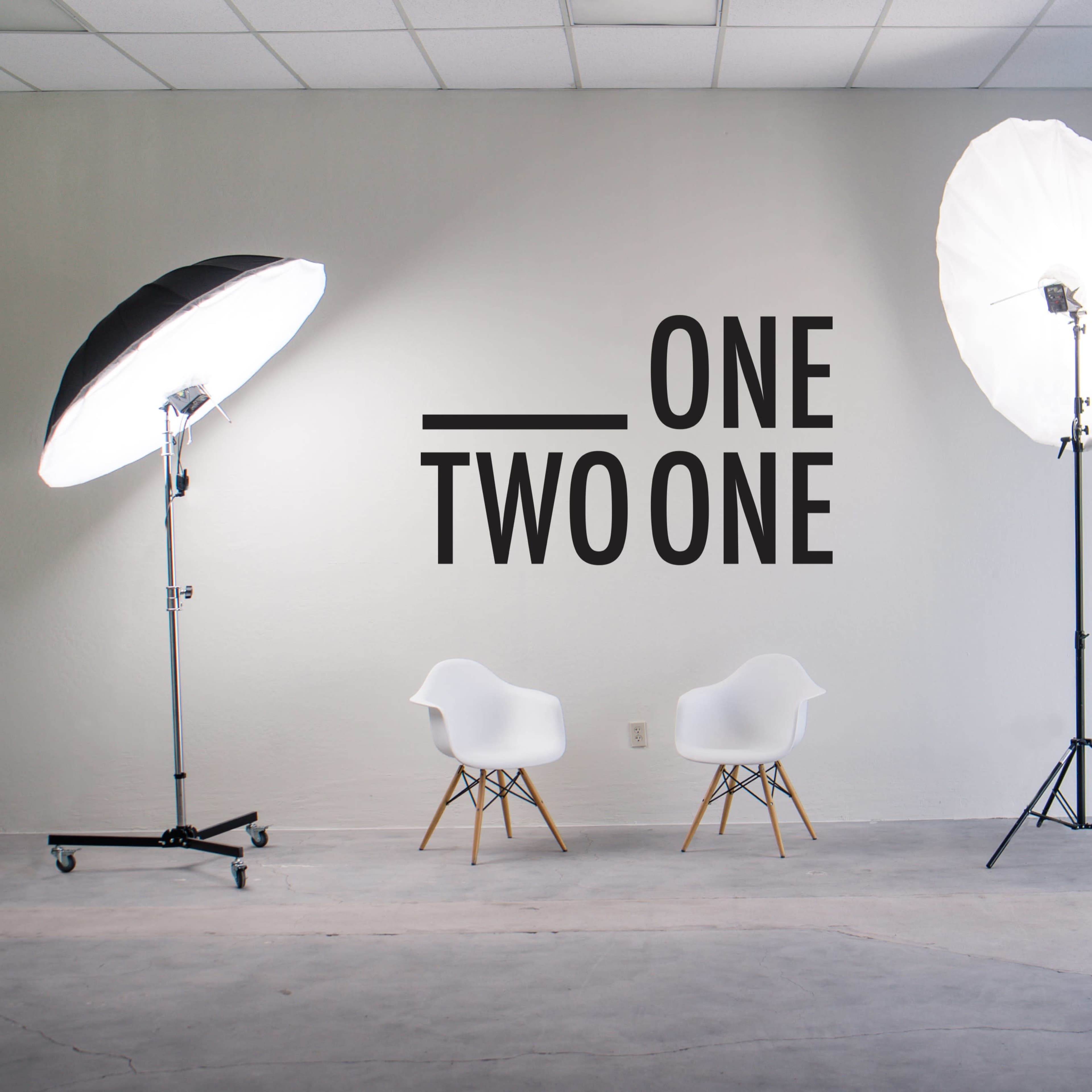 A photography studio with two softbox lights, two white chairs, and a wall featuring the text "ONE TWO ONE."