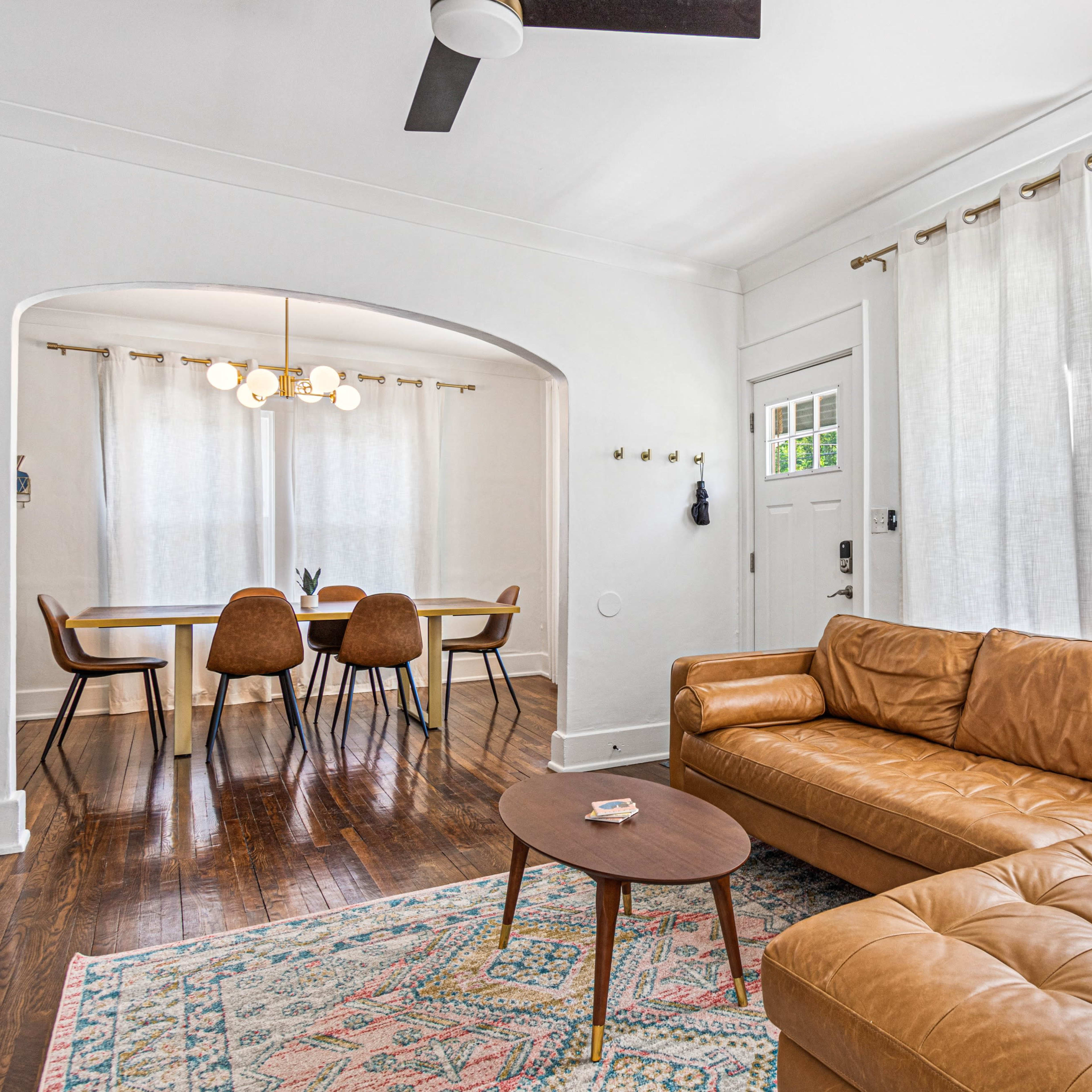 A modern living room features a brown leather sofa, a round coffee table, and light-colored curtains, with a dining area visible in the background.