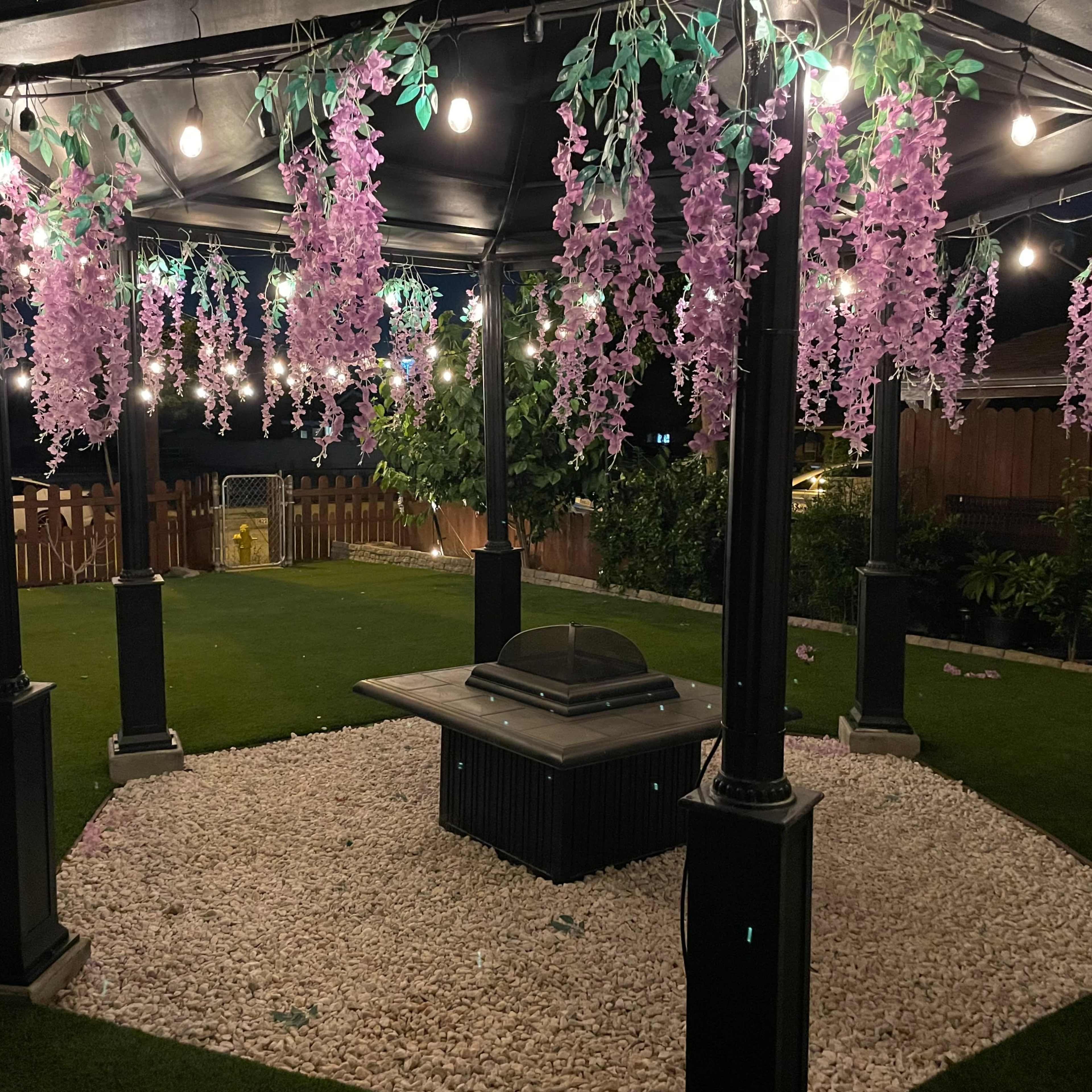 A gazebo adorned with purple flowers and string lights sits on a grassy area surrounded by decorative gravel.