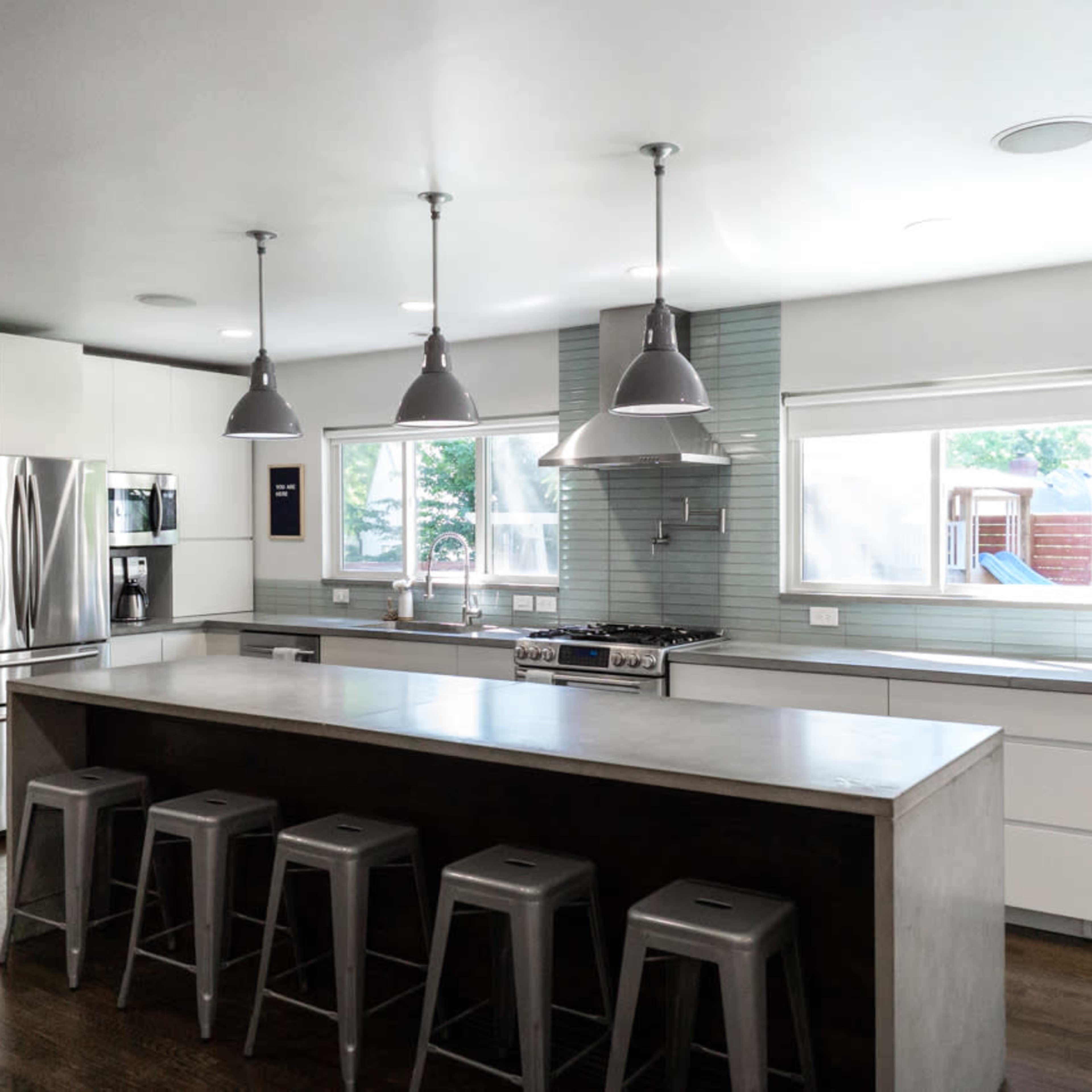 The modern kitchen features a central concrete island with metal stools, stainless steel appliances, and large windows that allow natural light to fill the space.