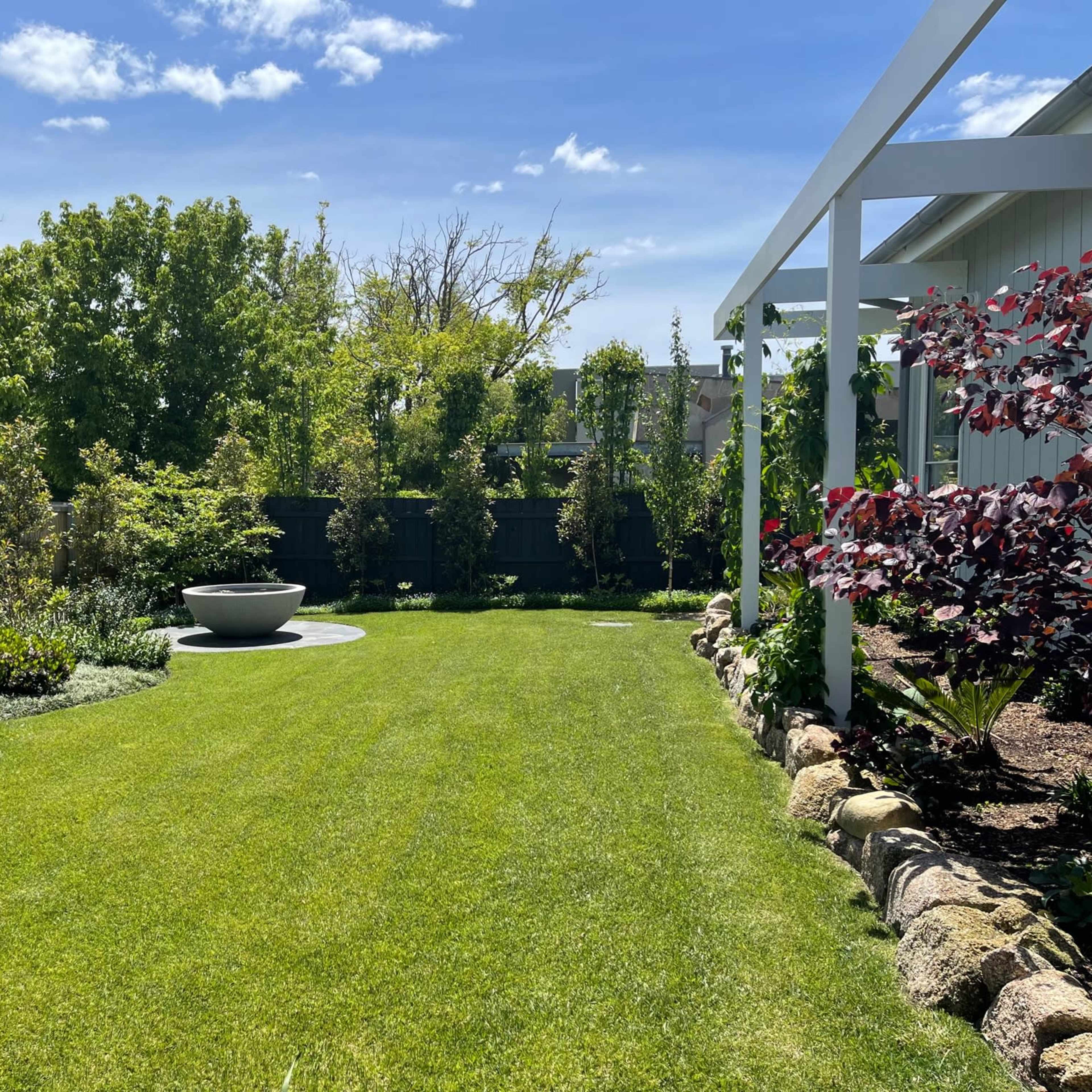 A landscaped garden with a green lawn, stone pathways, a circular seating area, and lush trees and shrubs.