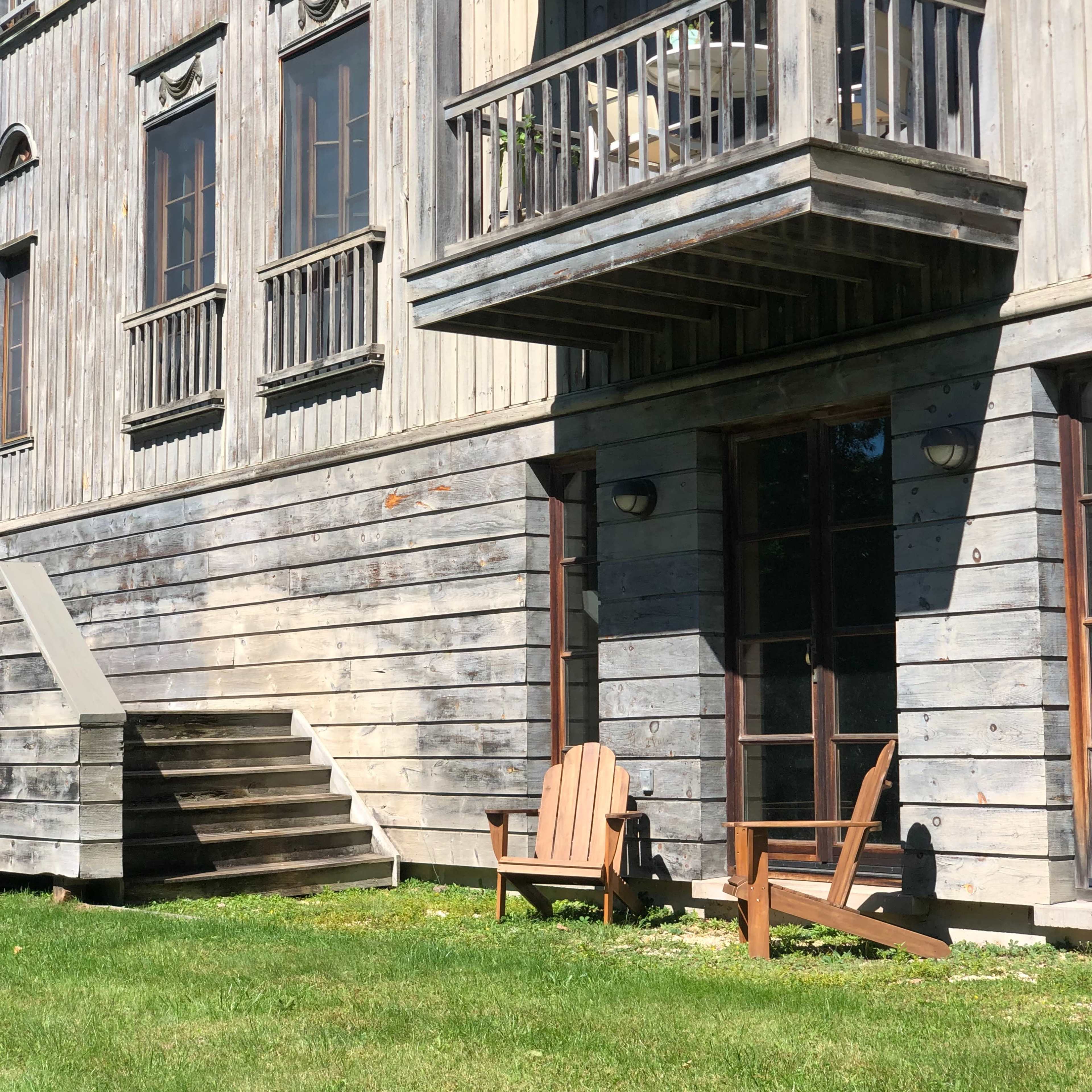 A wooden building with a balcony and a set of stairs features a pair of Adirondack chairs on the grass in front.