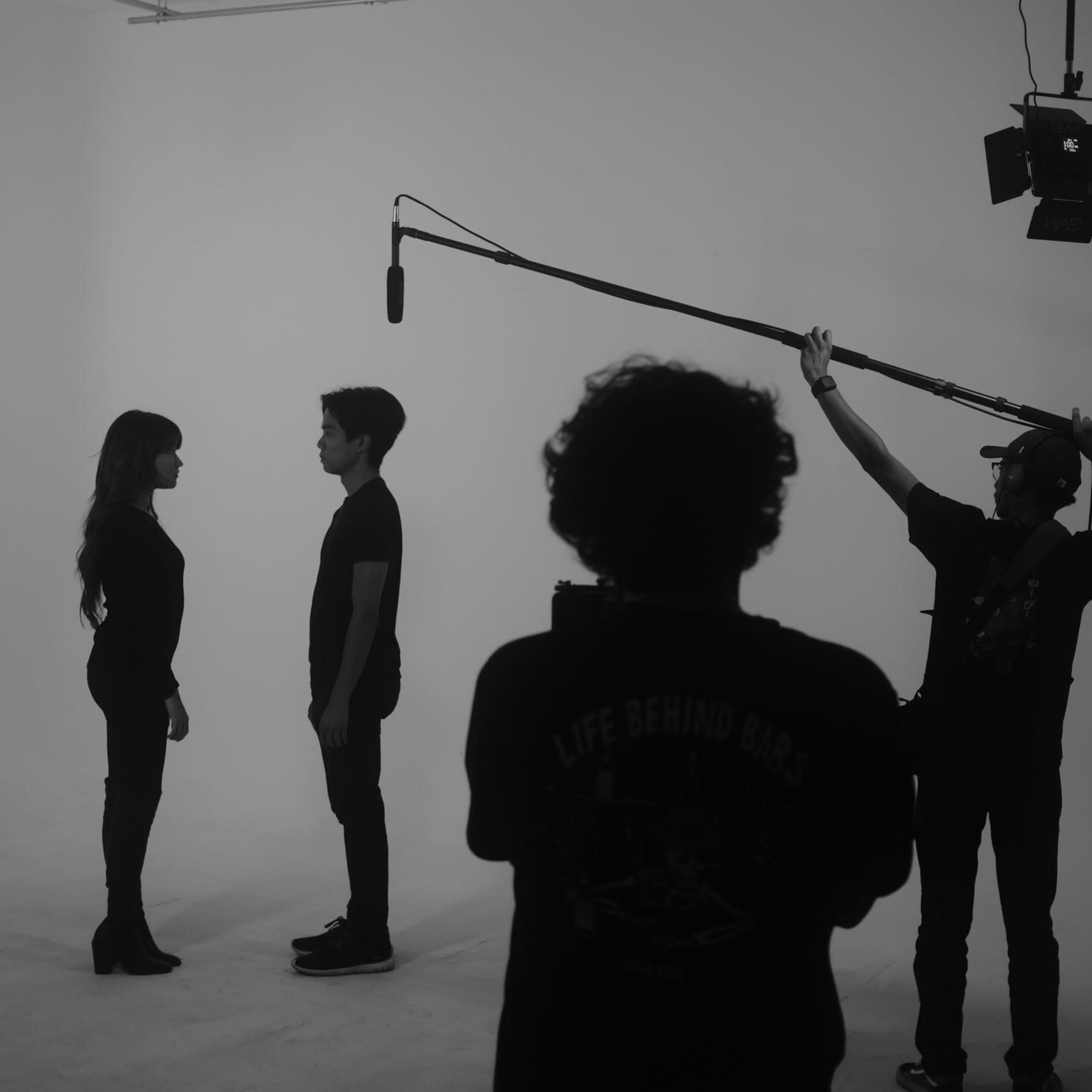 Two individuals stand facing each other in a studio setting, while a crew member holds a microphone on a boom.