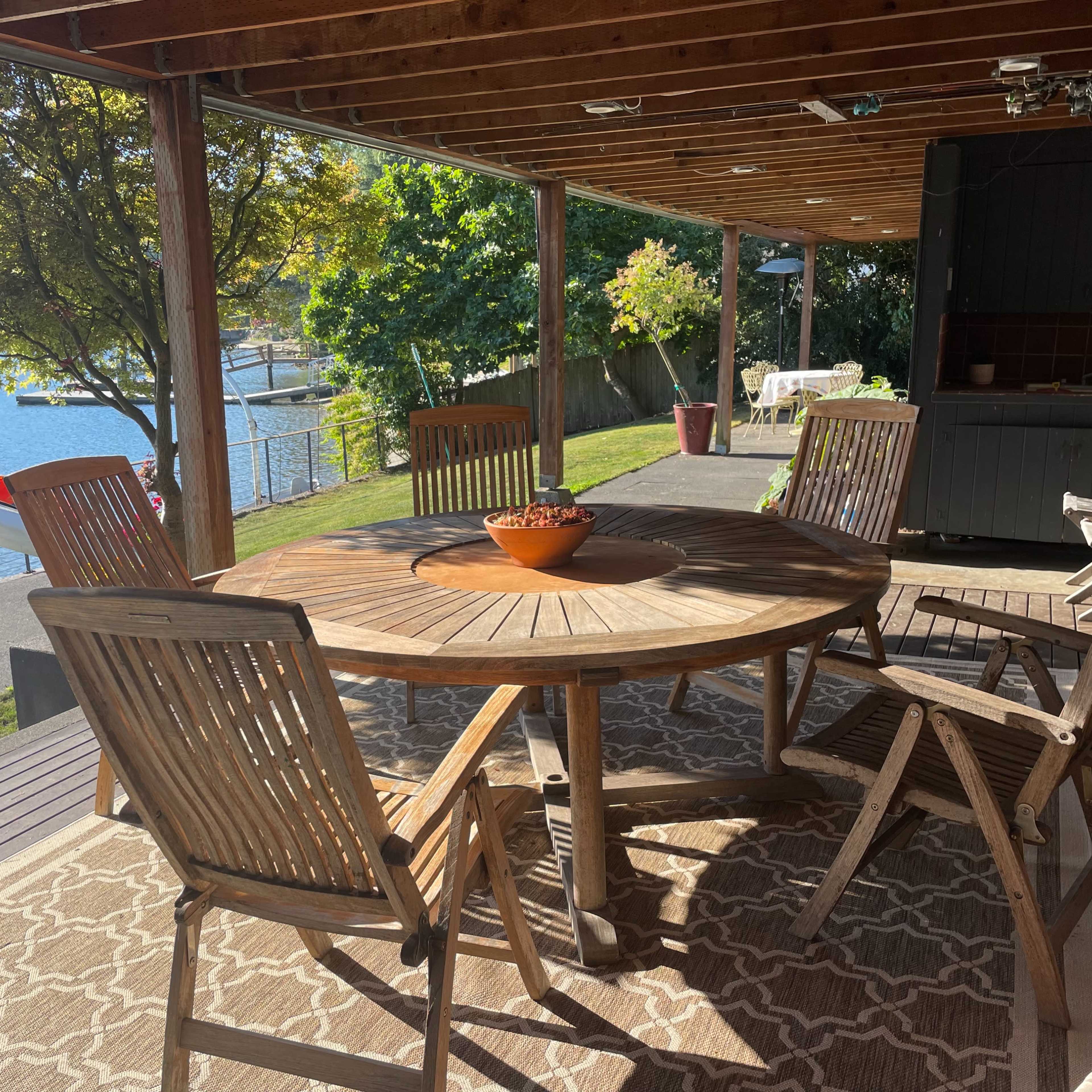 A wooden dining table with six chairs is set on a deck overlooking a lake, surrounded by greenery and a kayak nearby.