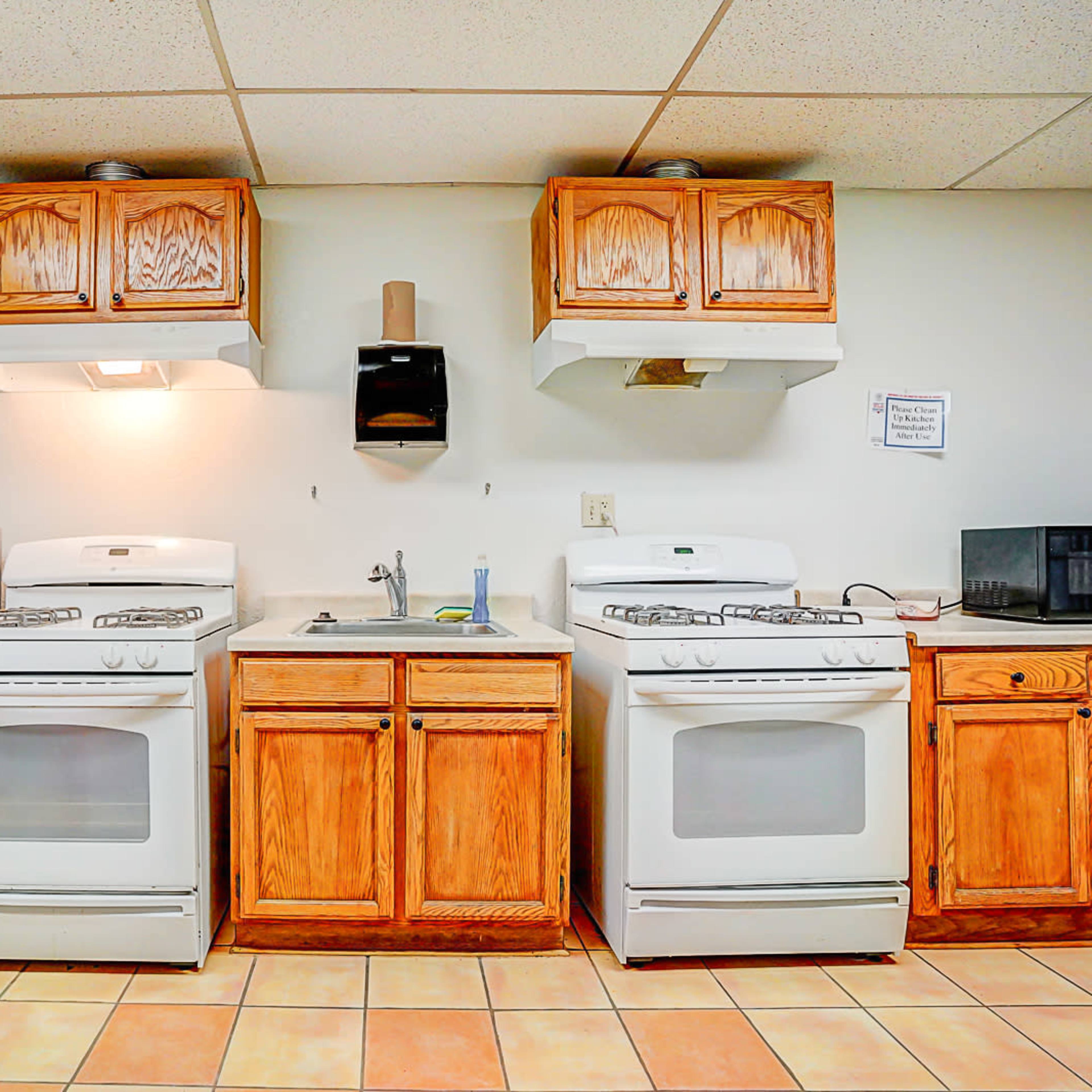 A kitchen with two white gas stoves, wooden cabinets, a stainless steel refrigerator, a microwave, and a sink between them.