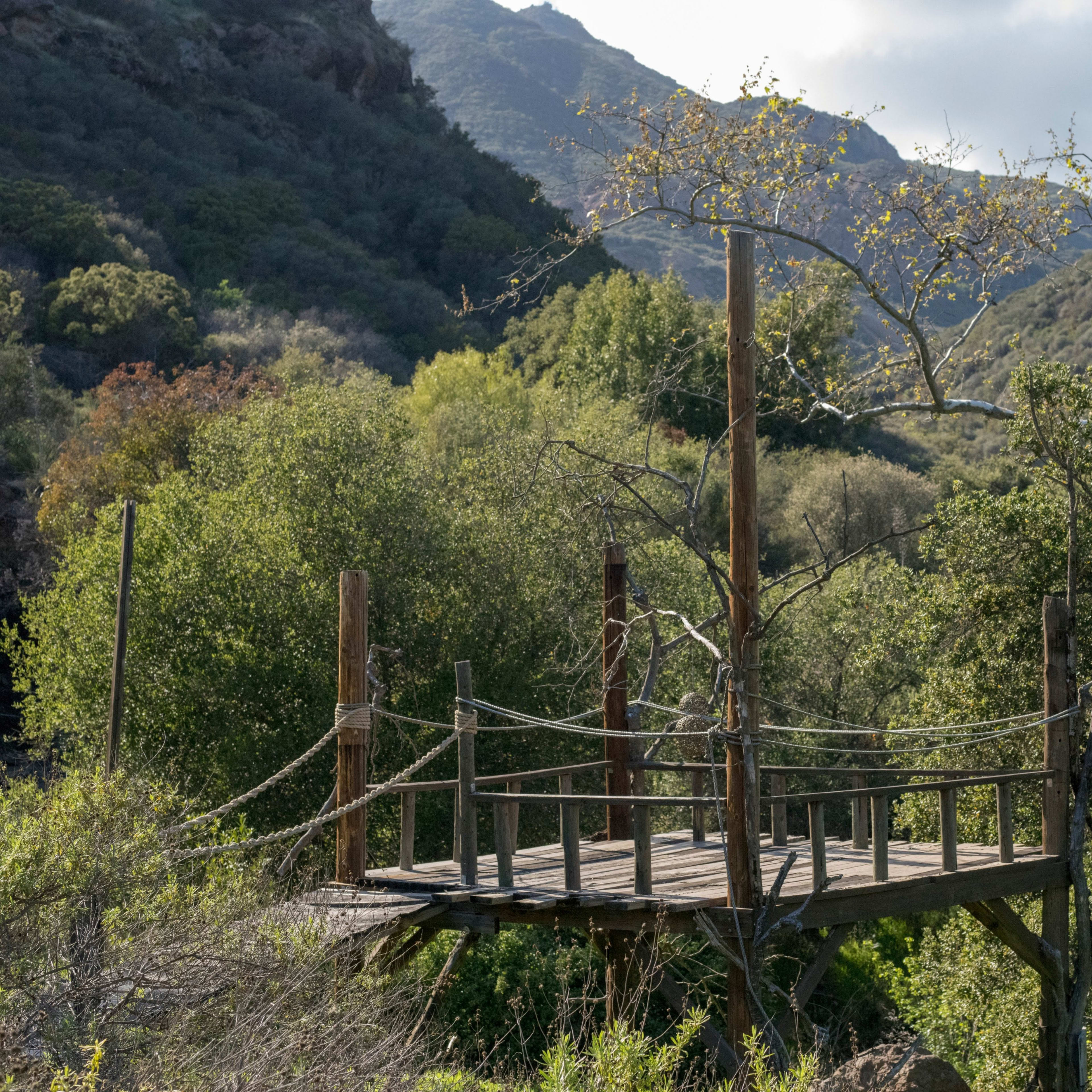 A wooden bridge supported by posts spans a wooded area surrounded by hills.