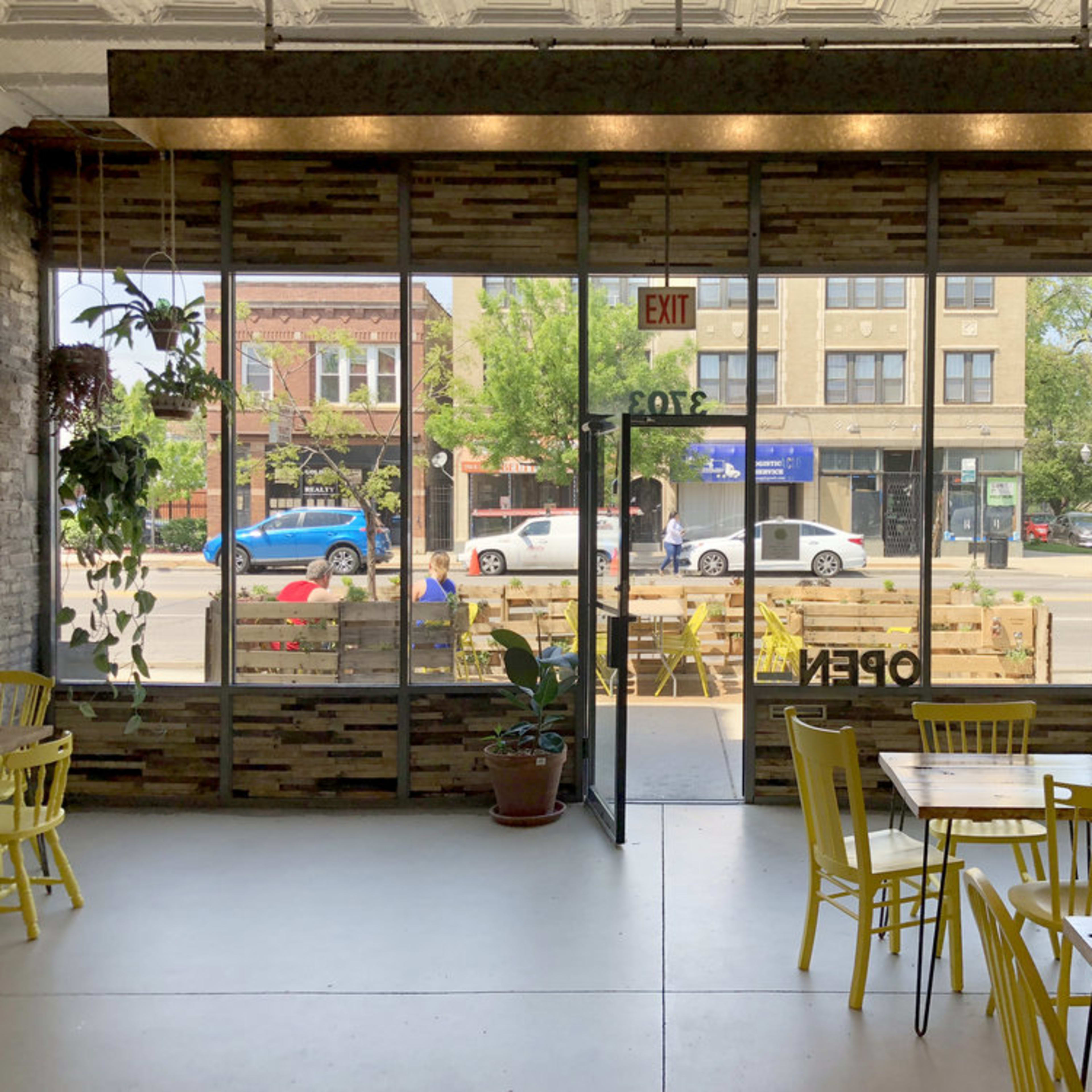 A bright café features large windows with a view of the street, wooden furniture, and plants hanging from the ceiling.
