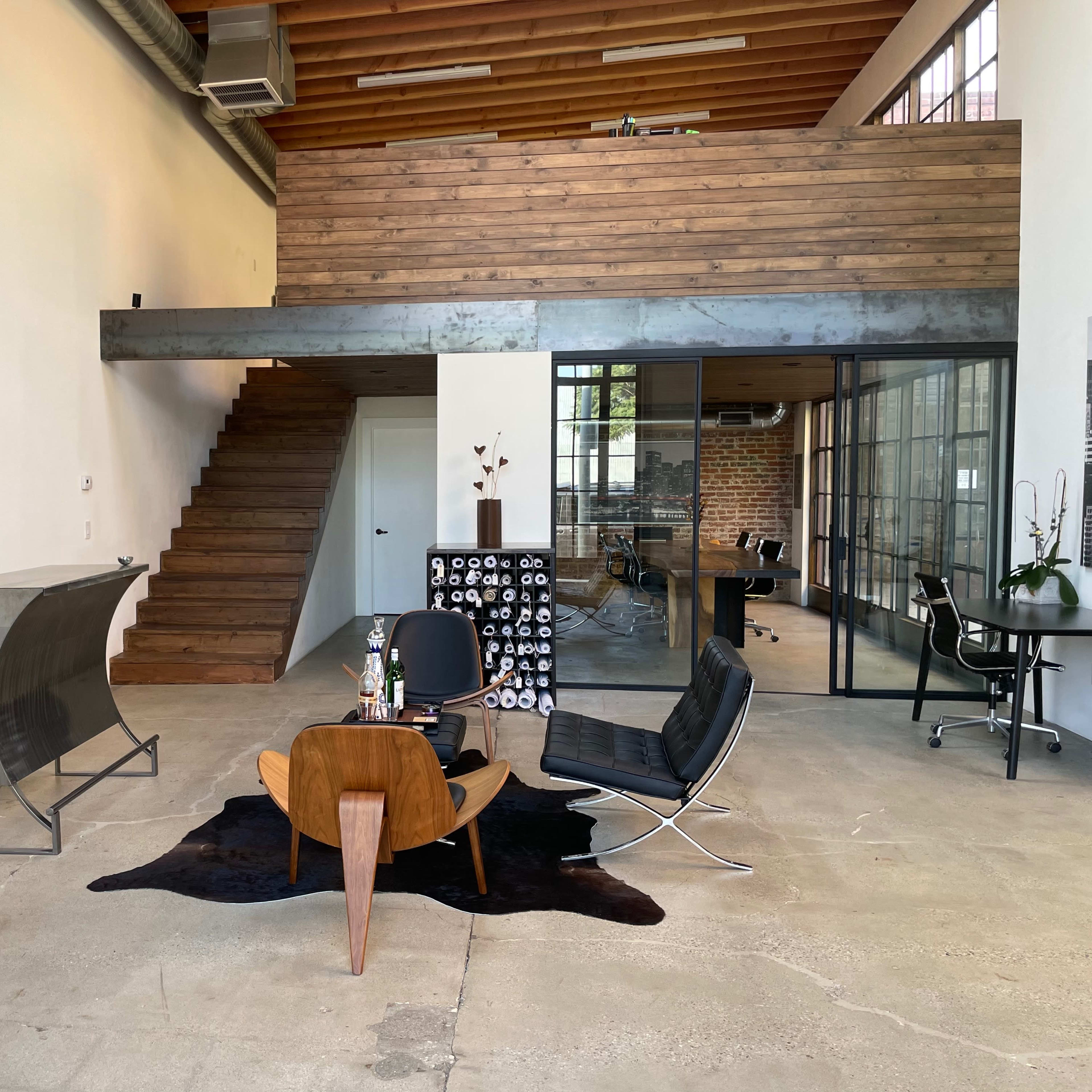 A spacious, modern office interior featuring a wooden staircase, a seating area with a coffee table and chairs, and a glass-walled meeting room.