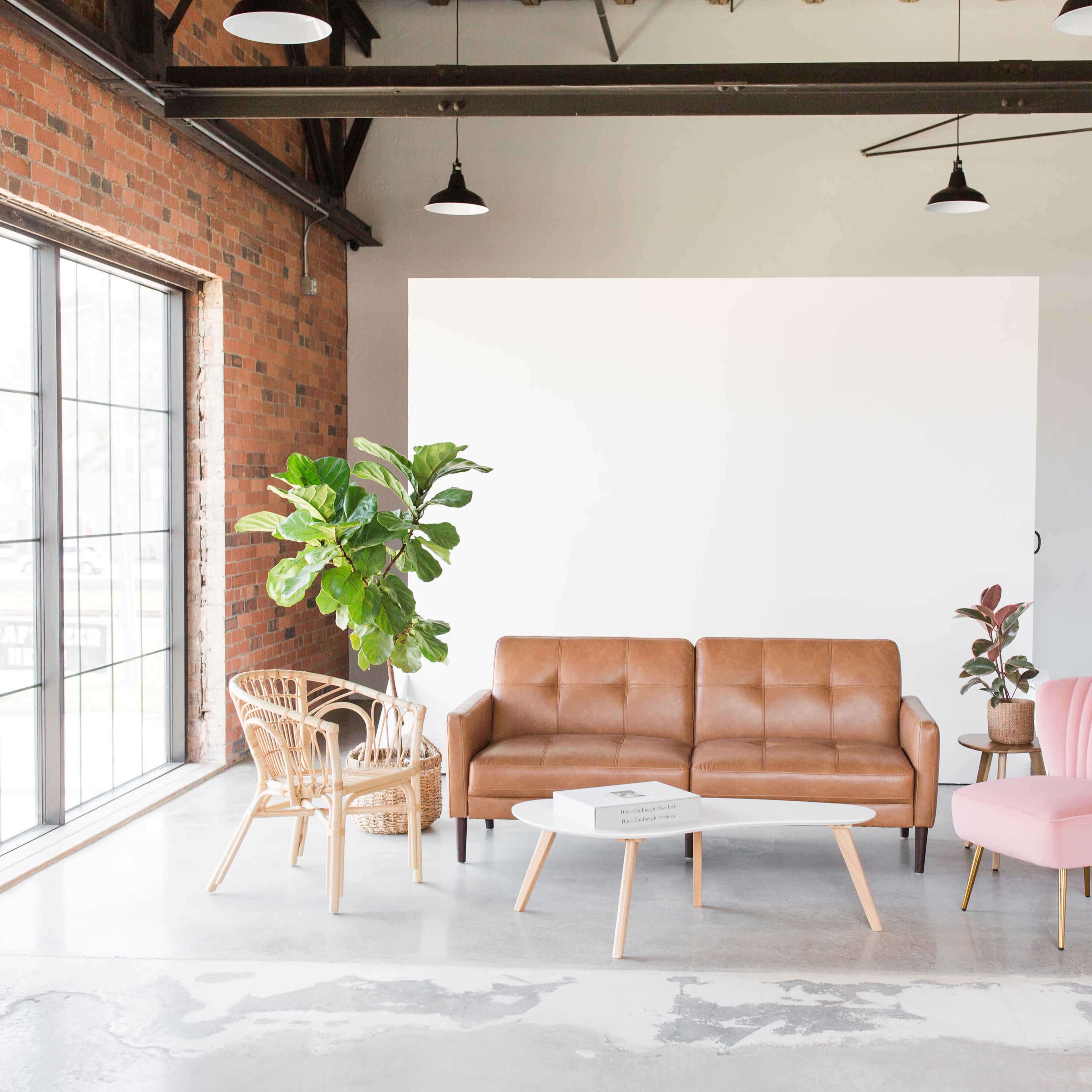 A modern interior space featuring a brown leather sofa, a pink armchair, a wooden chair, and a coffee table, complemented by greenery and an industrial-style brick wall.
