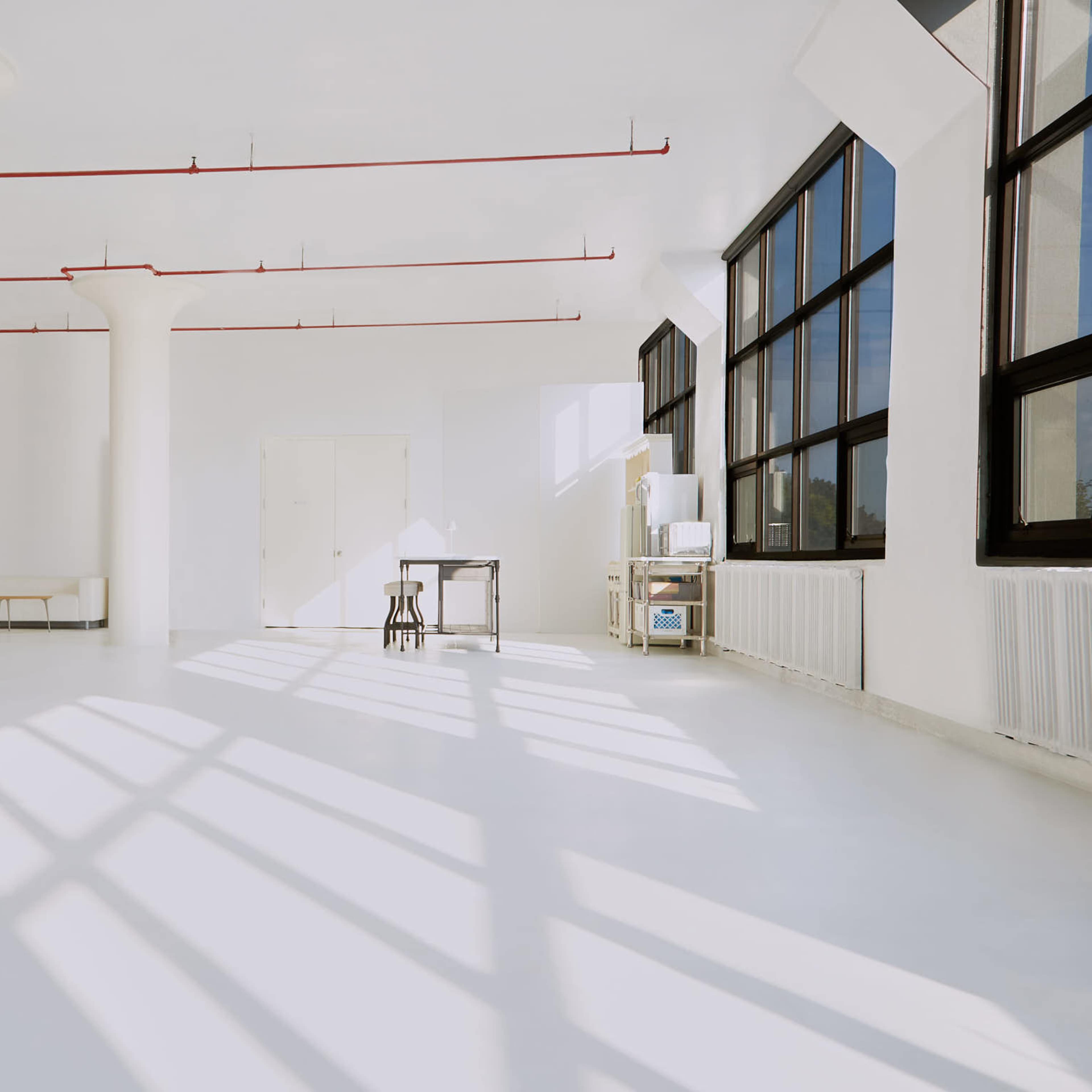 A spacious, bright room with white walls and floors, featuring large windows, a minimalistic table, and a few pillars.