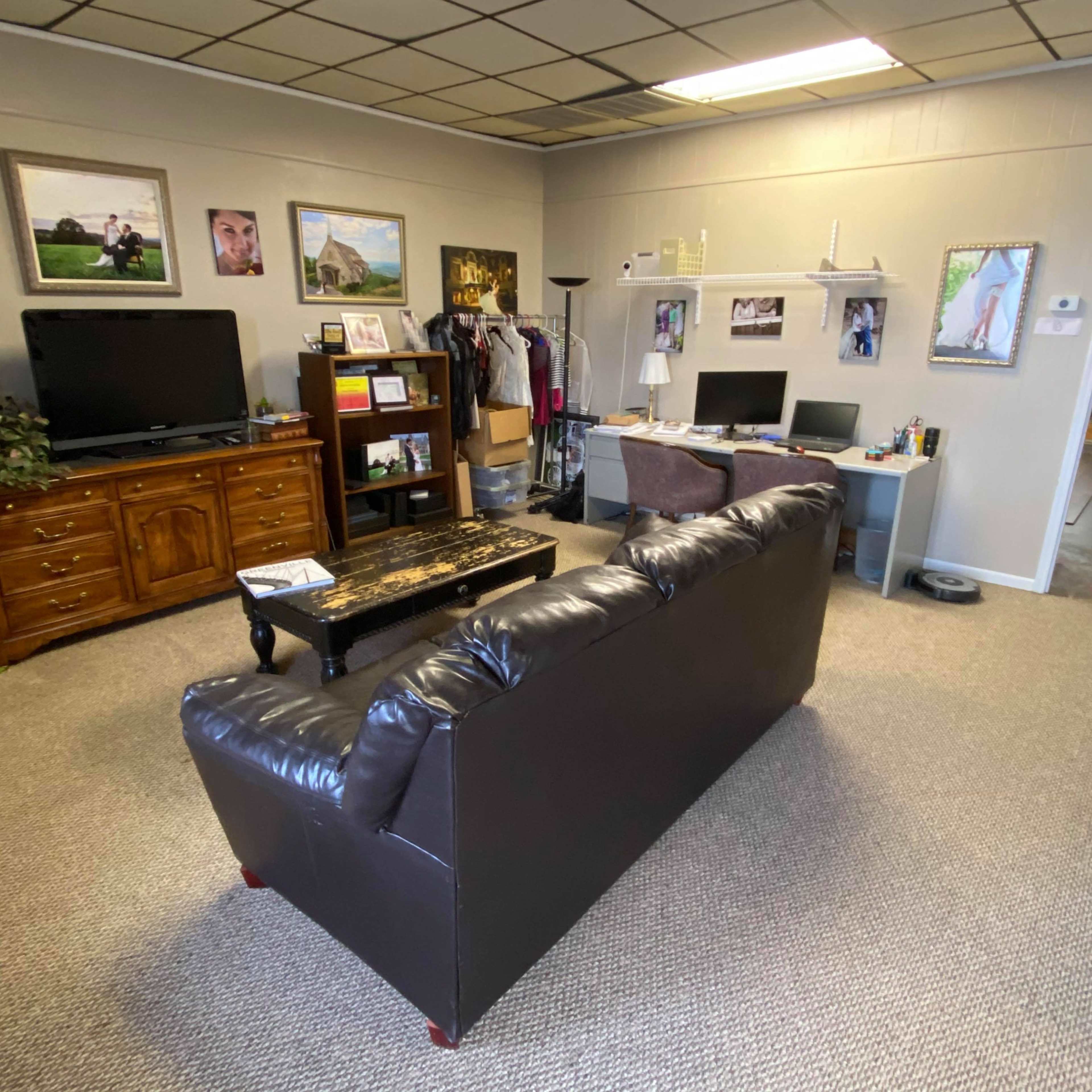 A small office space featuring a dark leather sofa, a wooden television cabinet, a desk with a computer, and framed photographs on the walls.