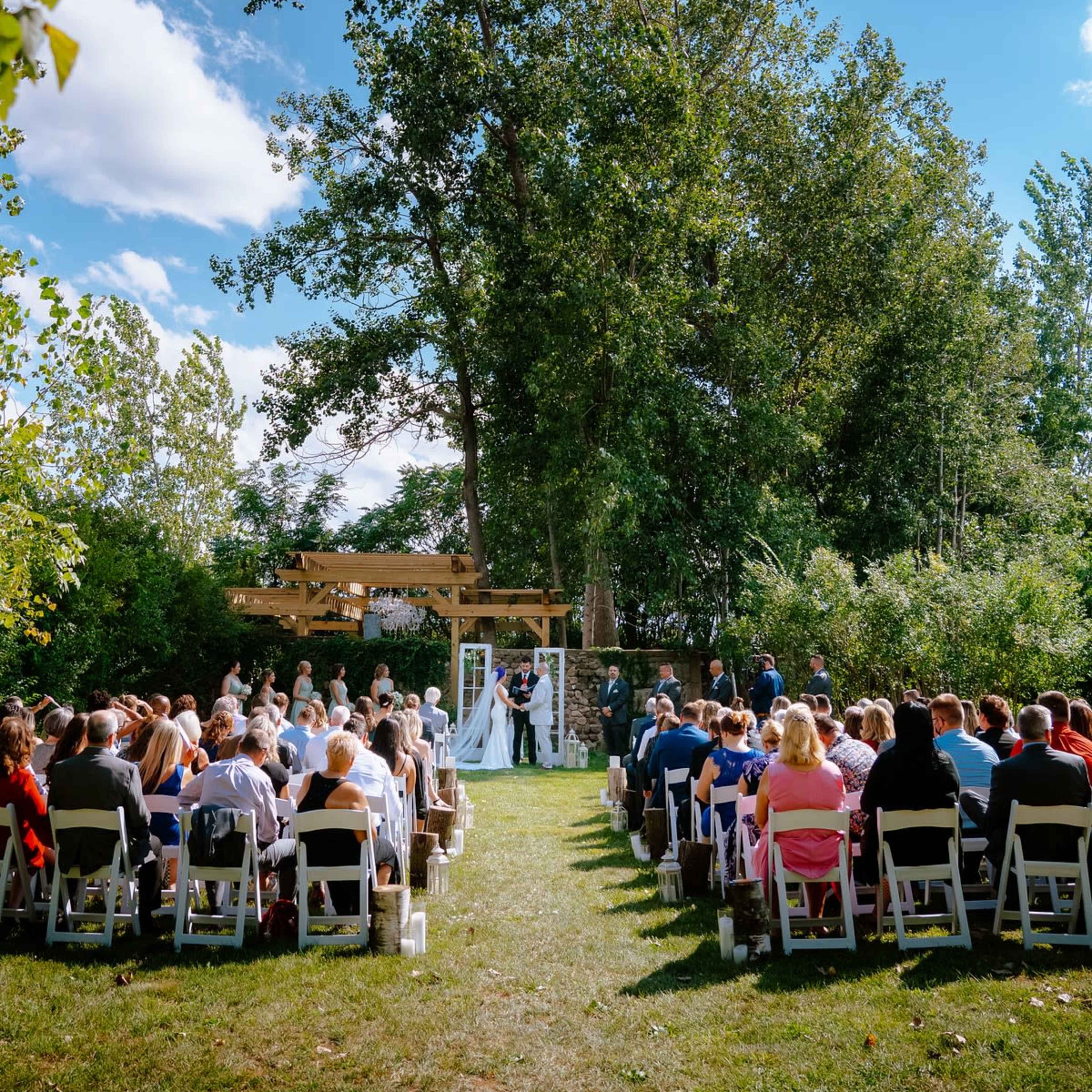 A wedding ceremony takes place outdoors under a wooden arch, surrounded by trees and seated guests.