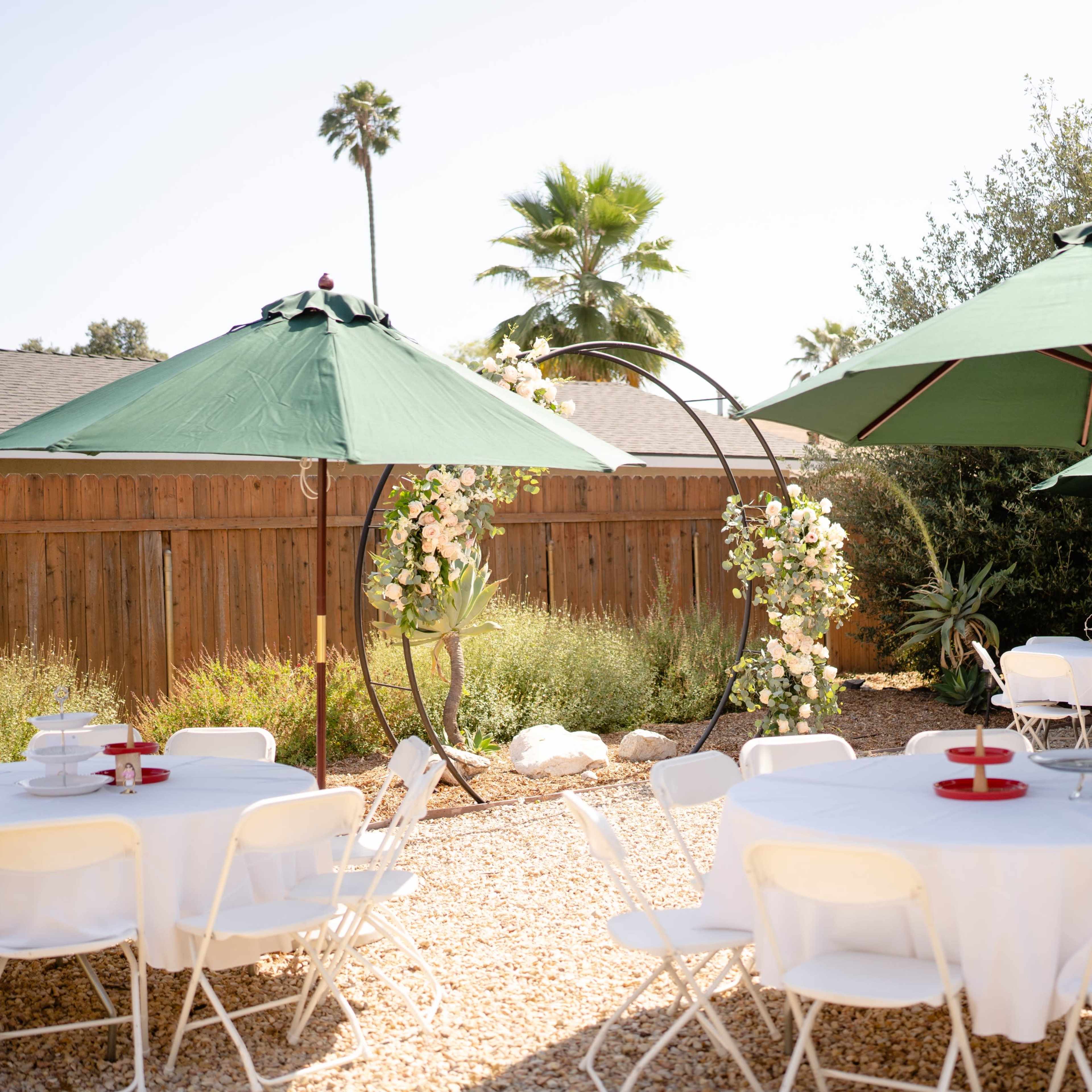 A backyard is set up for an outdoor gathering, featuring white tables and green umbrellas alongside greenery and a wooden fence.