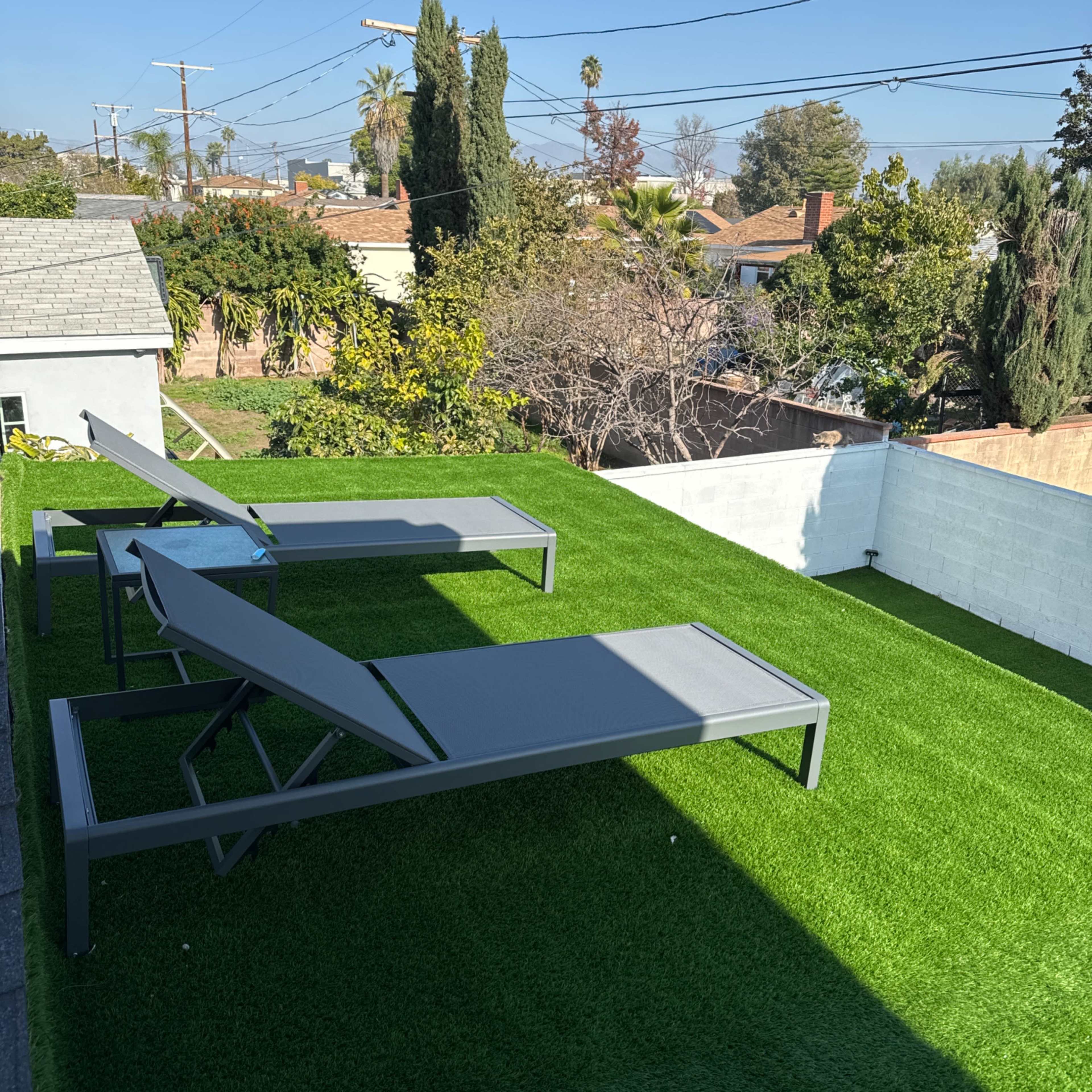 Two lounge chairs positioned on a green artificial lawn in a sunny backyard with trees and residential rooftops in the background.