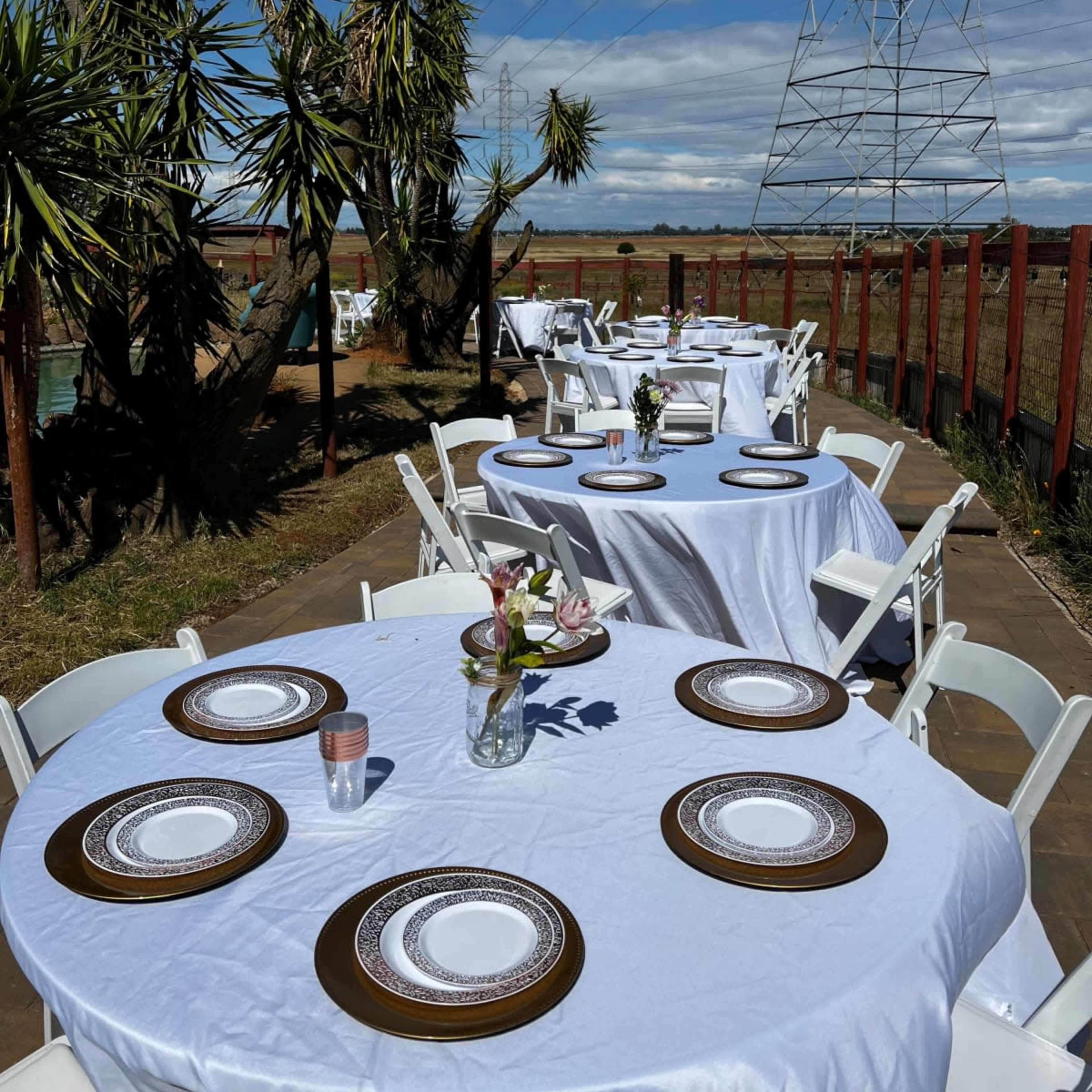 Several round tables covered with white tablecloths set up outdoors near a power line, with decorative centerpieces and chairs arranged around them.