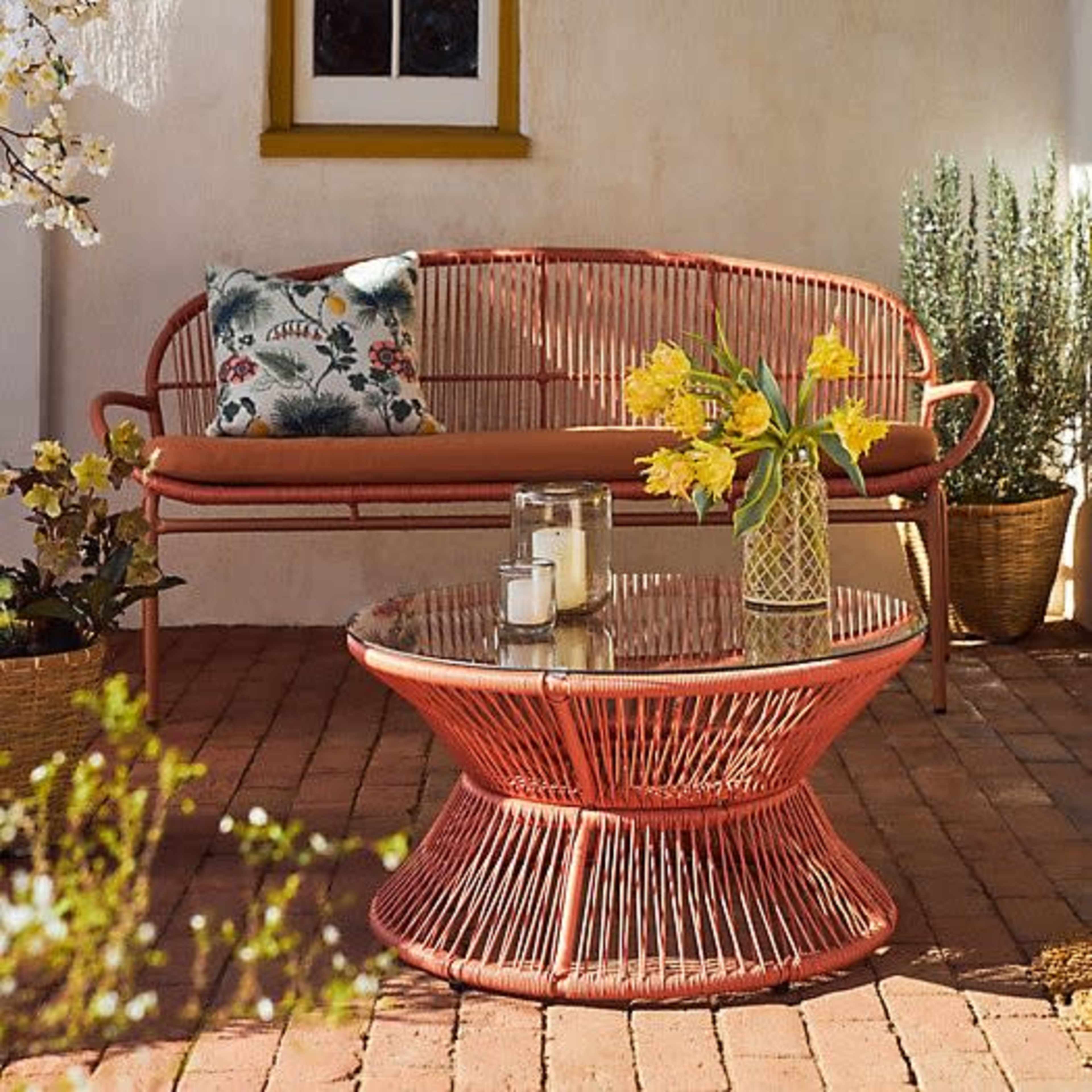 A wicker sofa and a round coffee table are placed on a brick patio, surrounded by potted plants and a vase of yellow flowers.