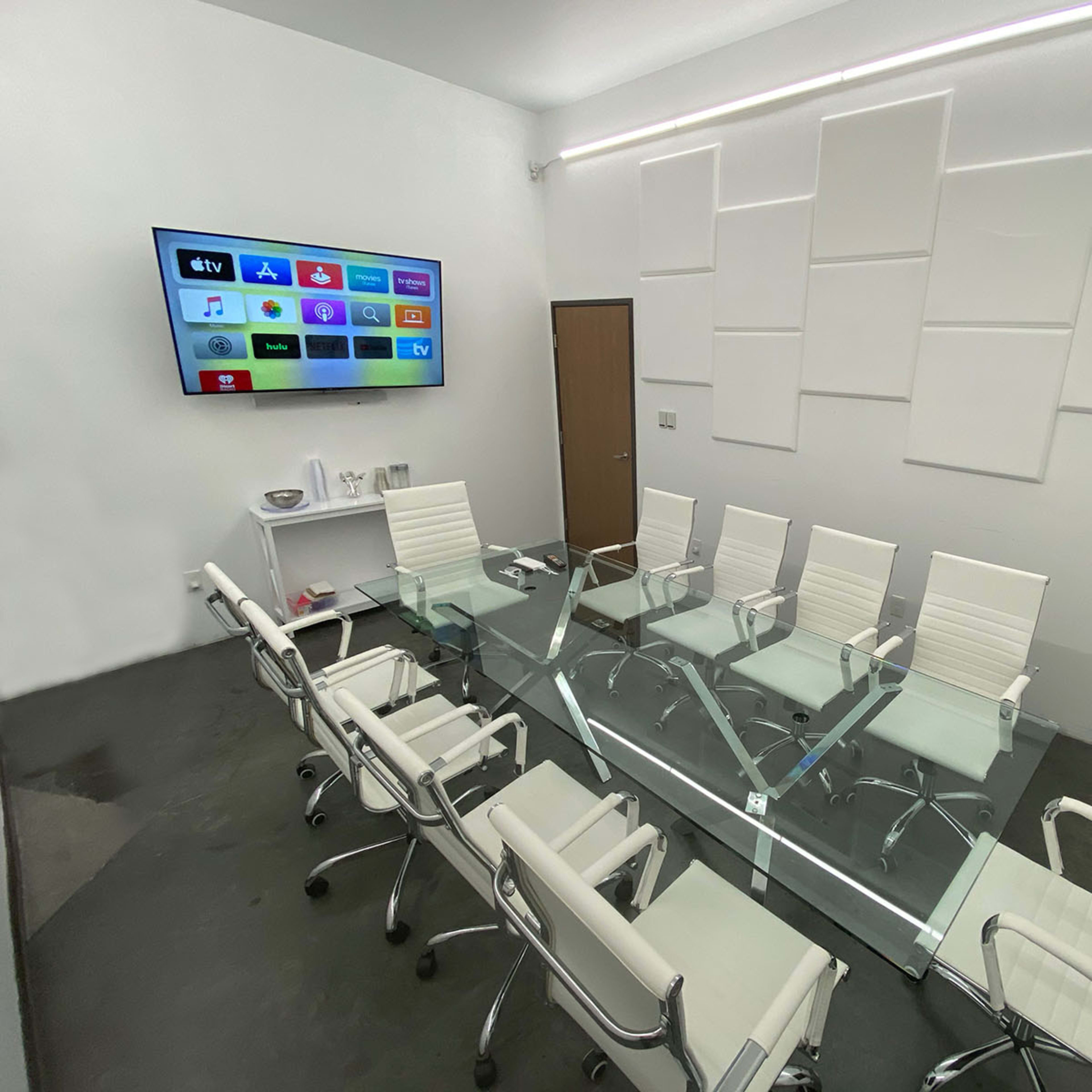 A modern conference room with a glass table surrounded by white chairs and a wall-mounted screen displaying various app icons.