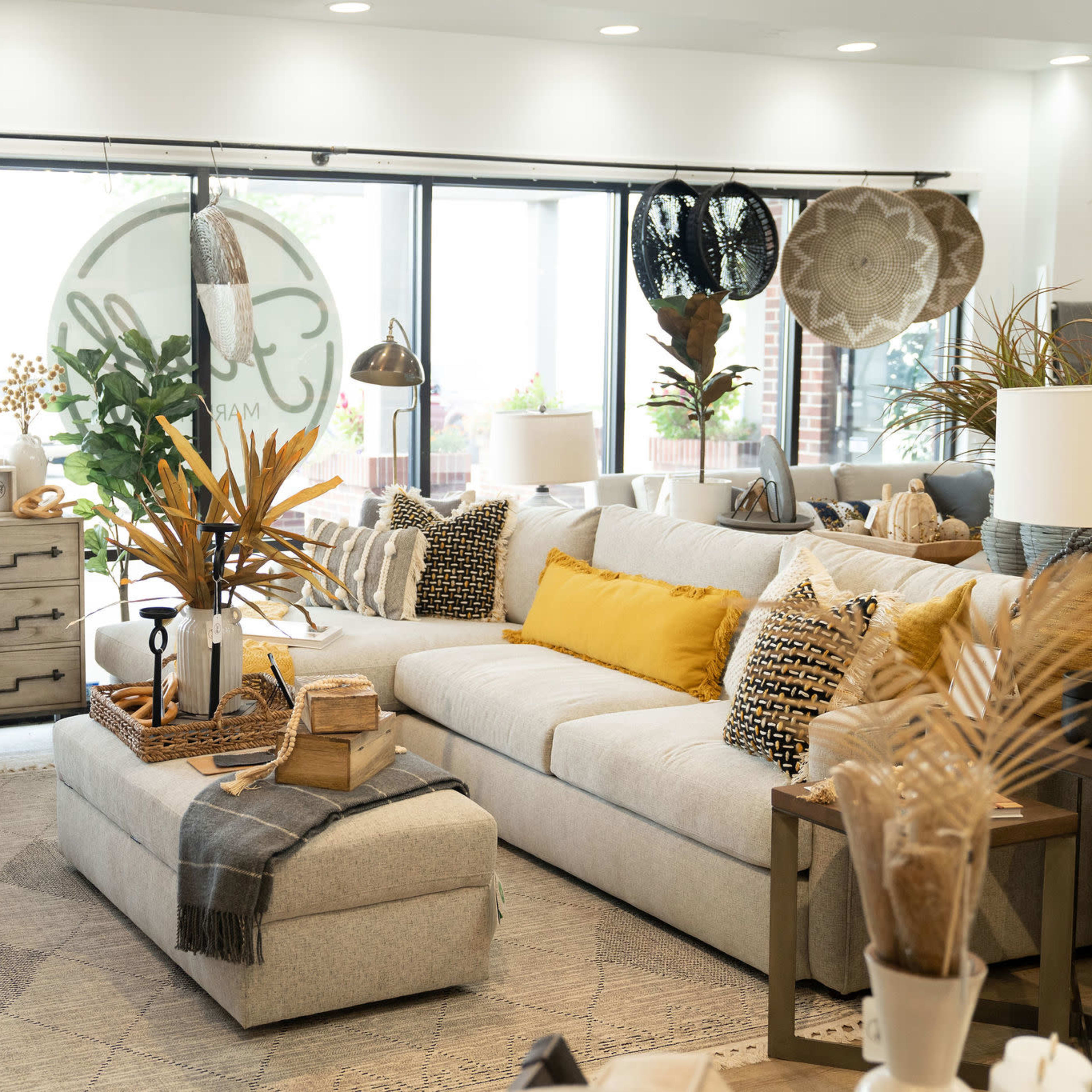 A modern living room features a spacious sectional couch with decorative pillows, a coffee table adorned with plants, and various textured furnishings beside large windows.