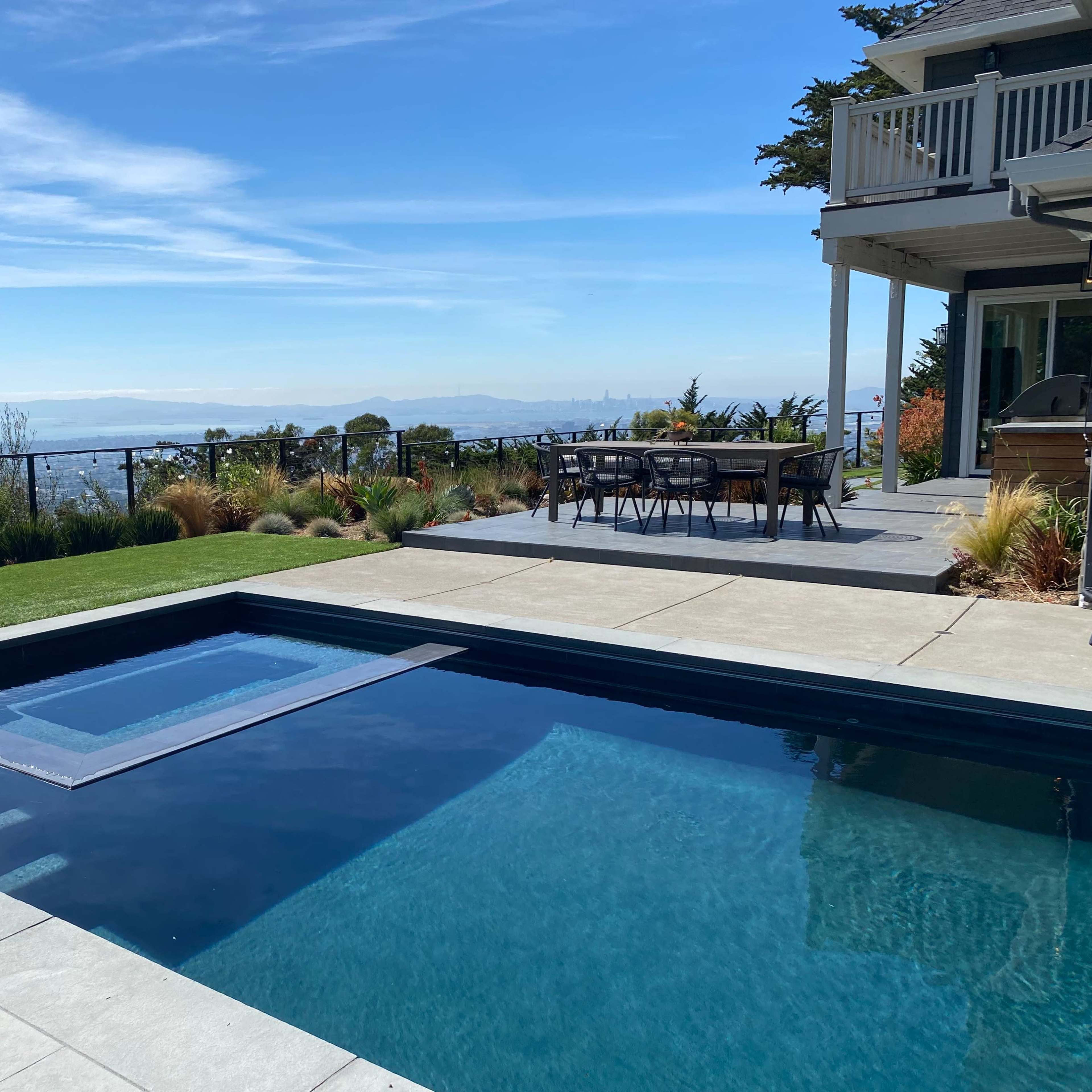 A rectangular swimming pool with a spa is situated next to a paved patio and grass lawn, overlooking a distant view of hills and a city skyline.