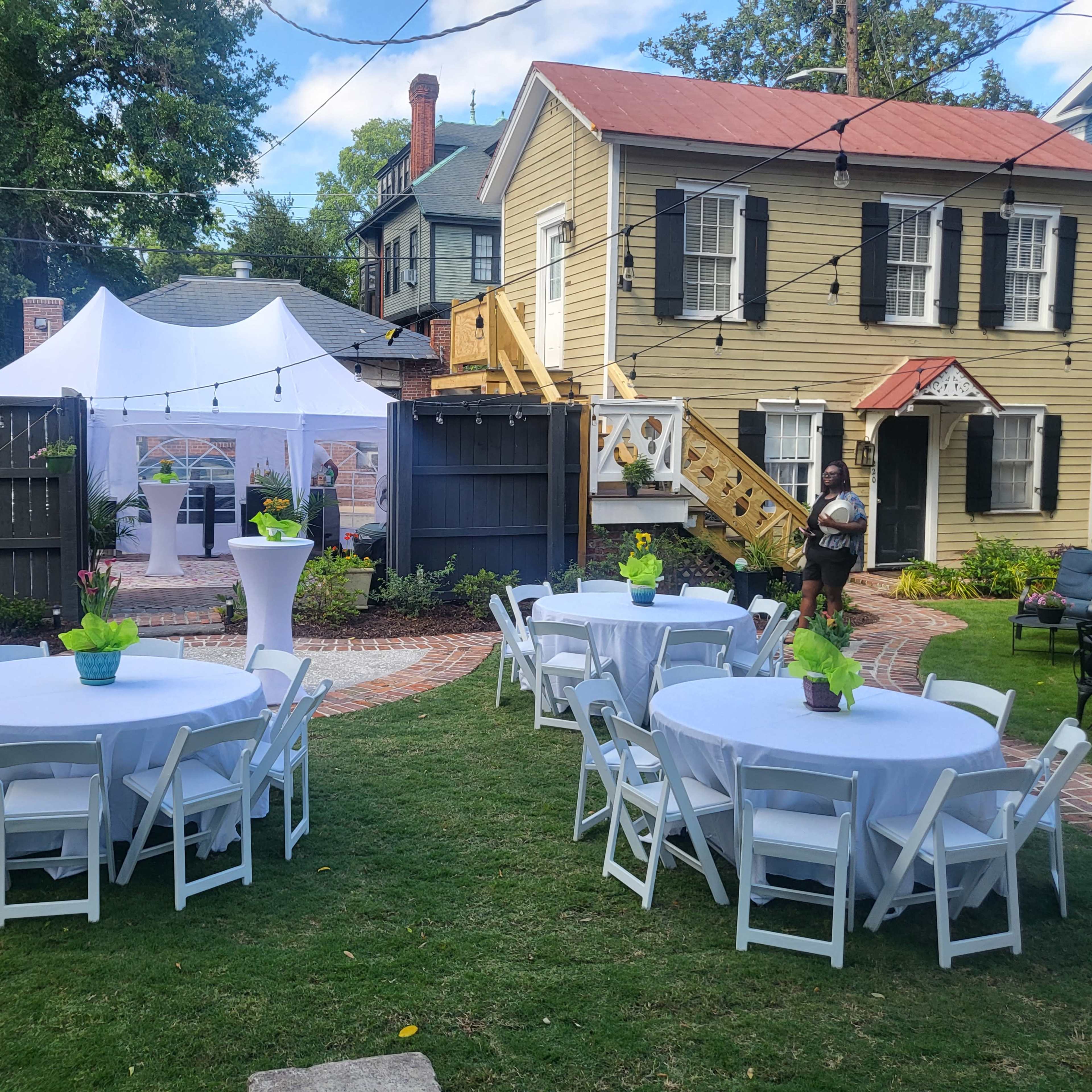 A courtyard setup for an event features round tables with white tablecloths and green centerpieces, surrounded by a white tent and a two-story house in the background.