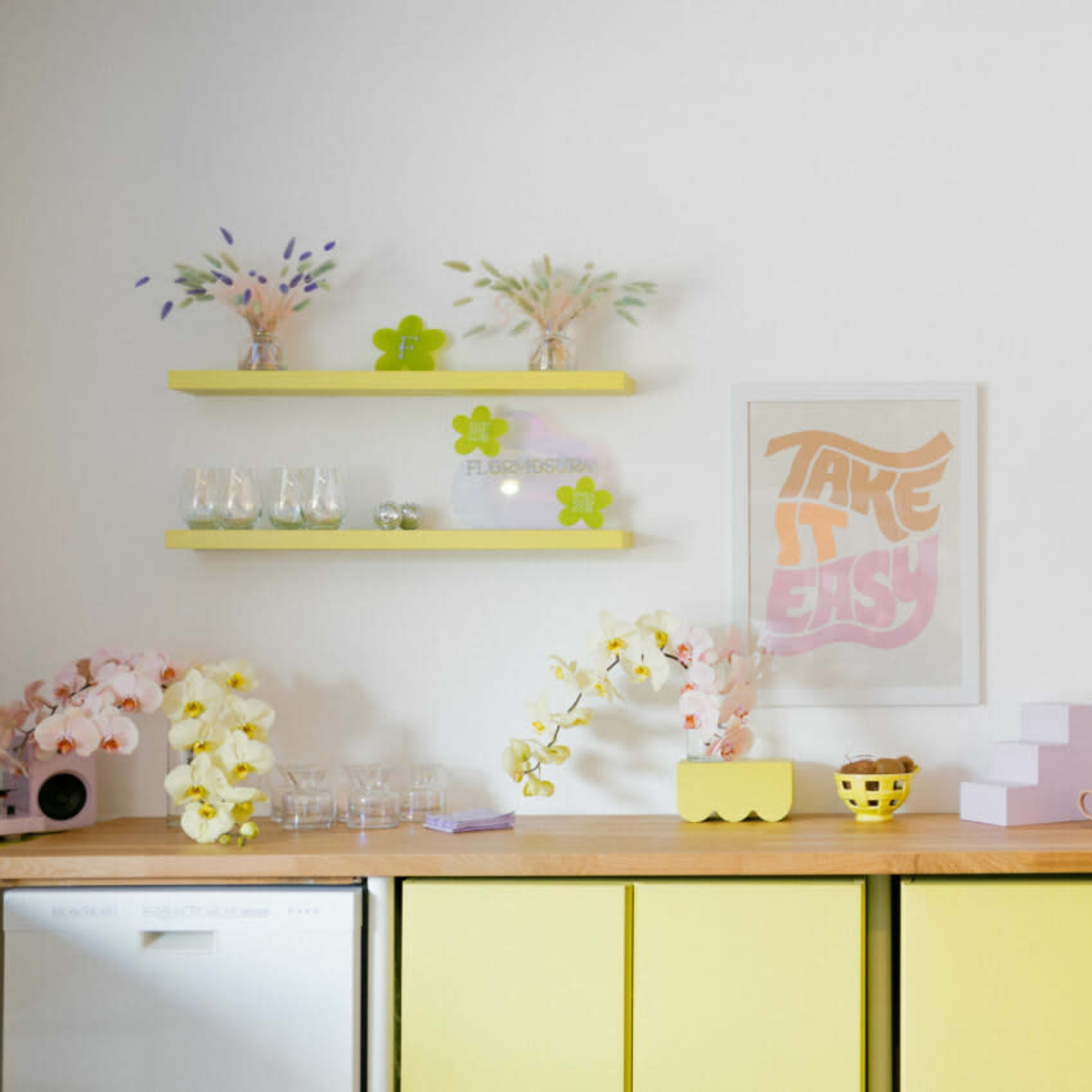 A bright kitchen countertop features yellow shelves with decorative items, a framed print, and various containers arranged neatly.