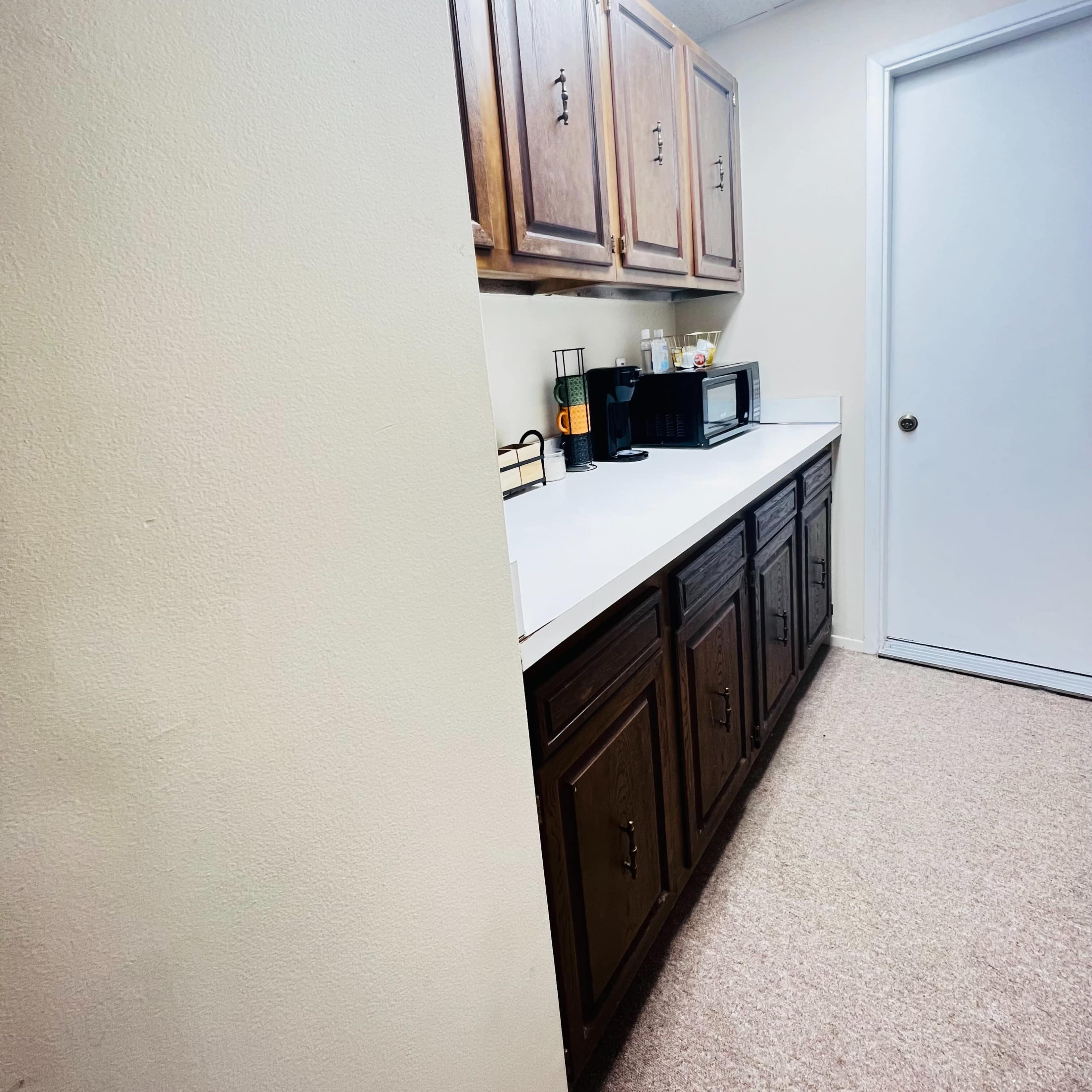 A narrow kitchen space with wooden cabinets above a white countertop and a microwave on the counter, leading to a closed door on the right.