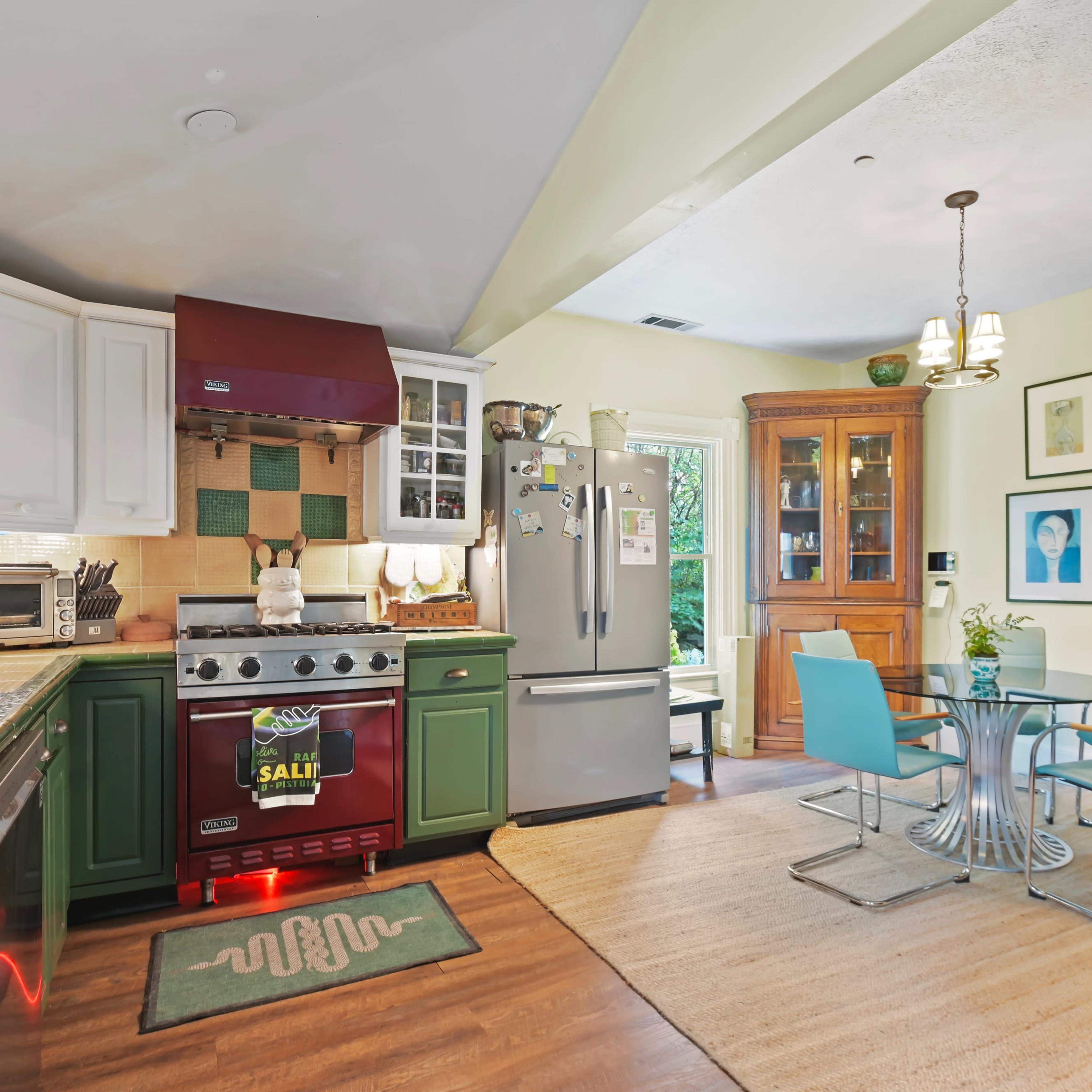 A kitchen featuring green cabinetry, stainless steel appliances, a circular dining table with blue chairs, and a wooden cabinet displaying framed artwork.