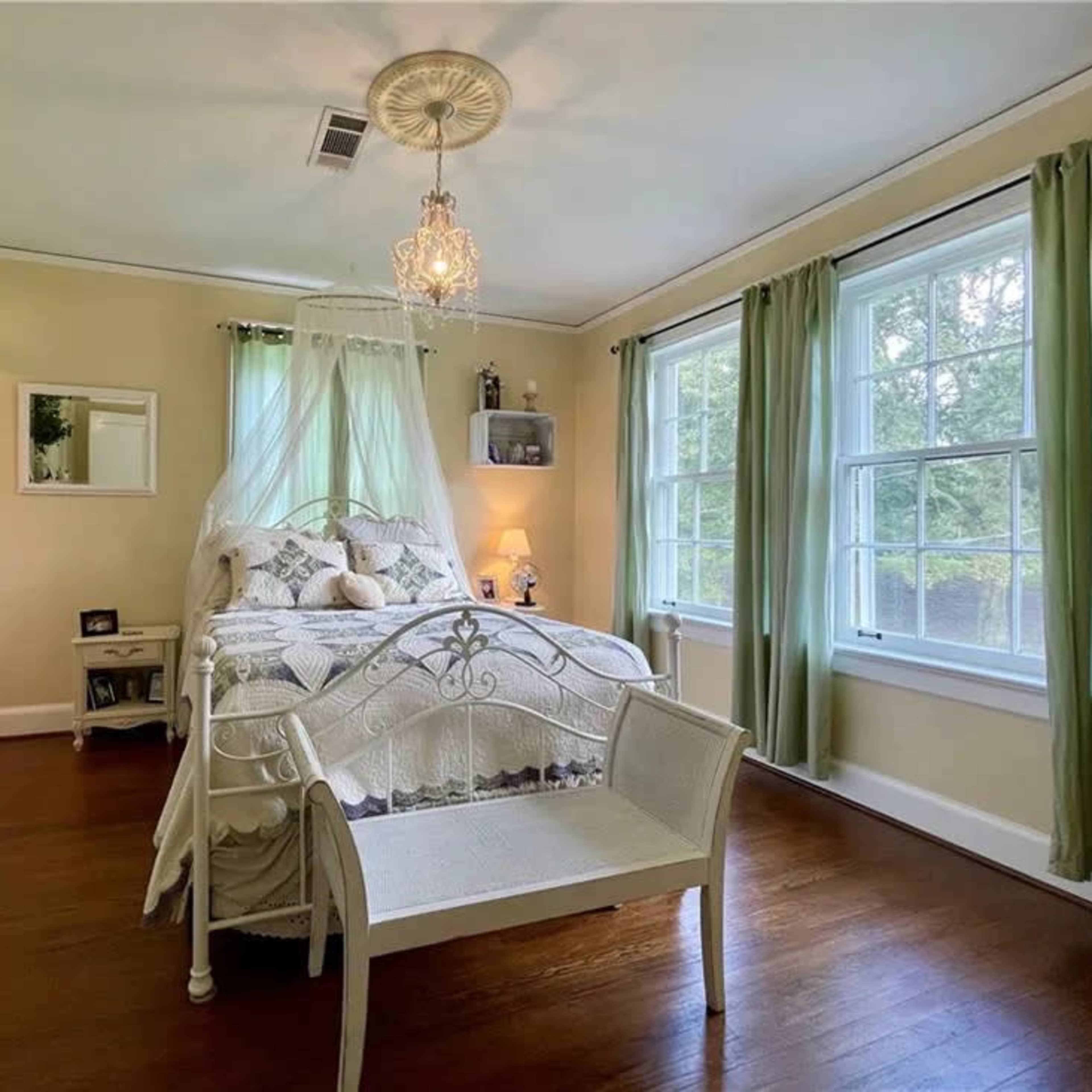 A cozy bedroom featuring a white bed with a canopy, a dresser, and large windows adorned with green curtains.