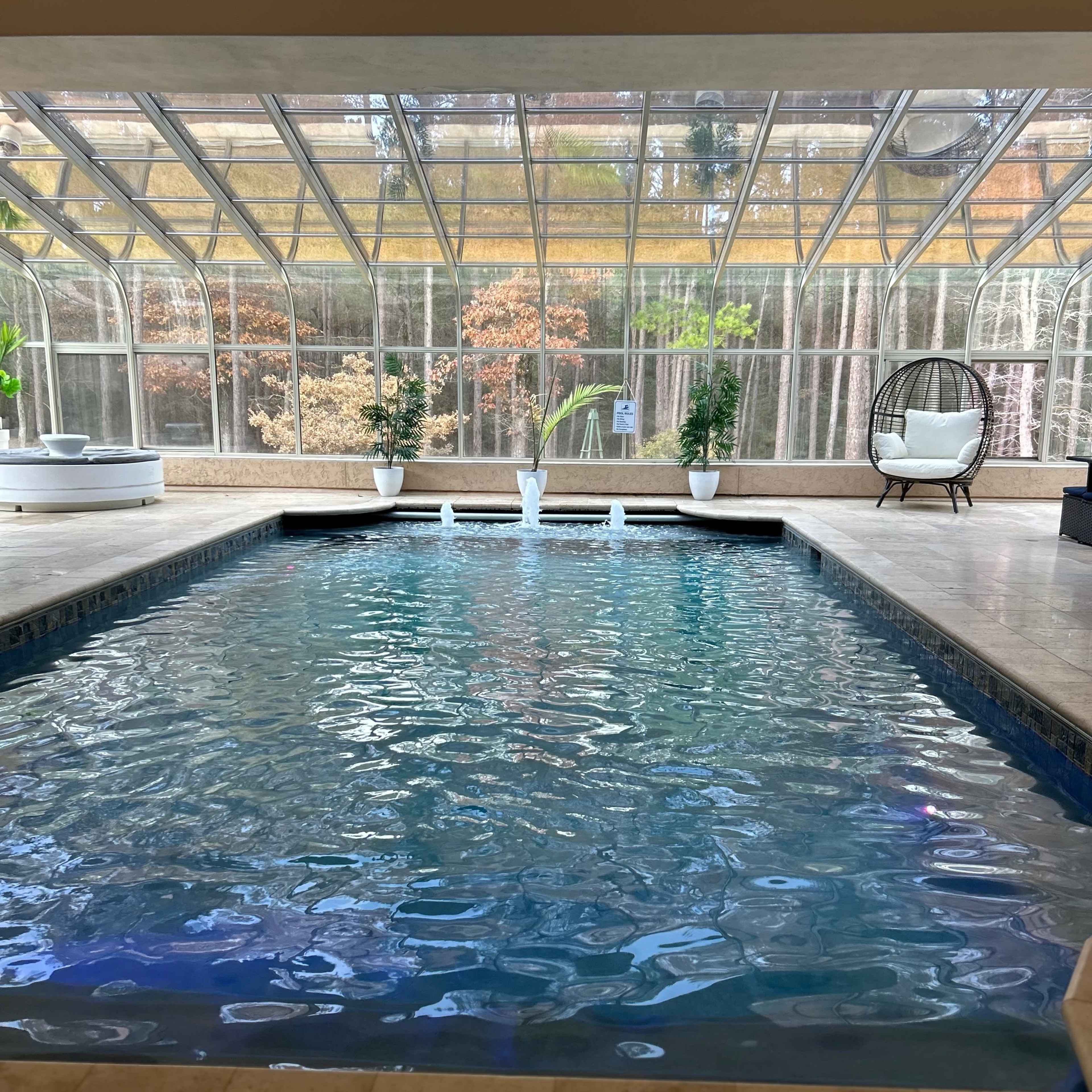 An indoor pool is framed by large glass panels, surrounded by potted plants and seating areas.