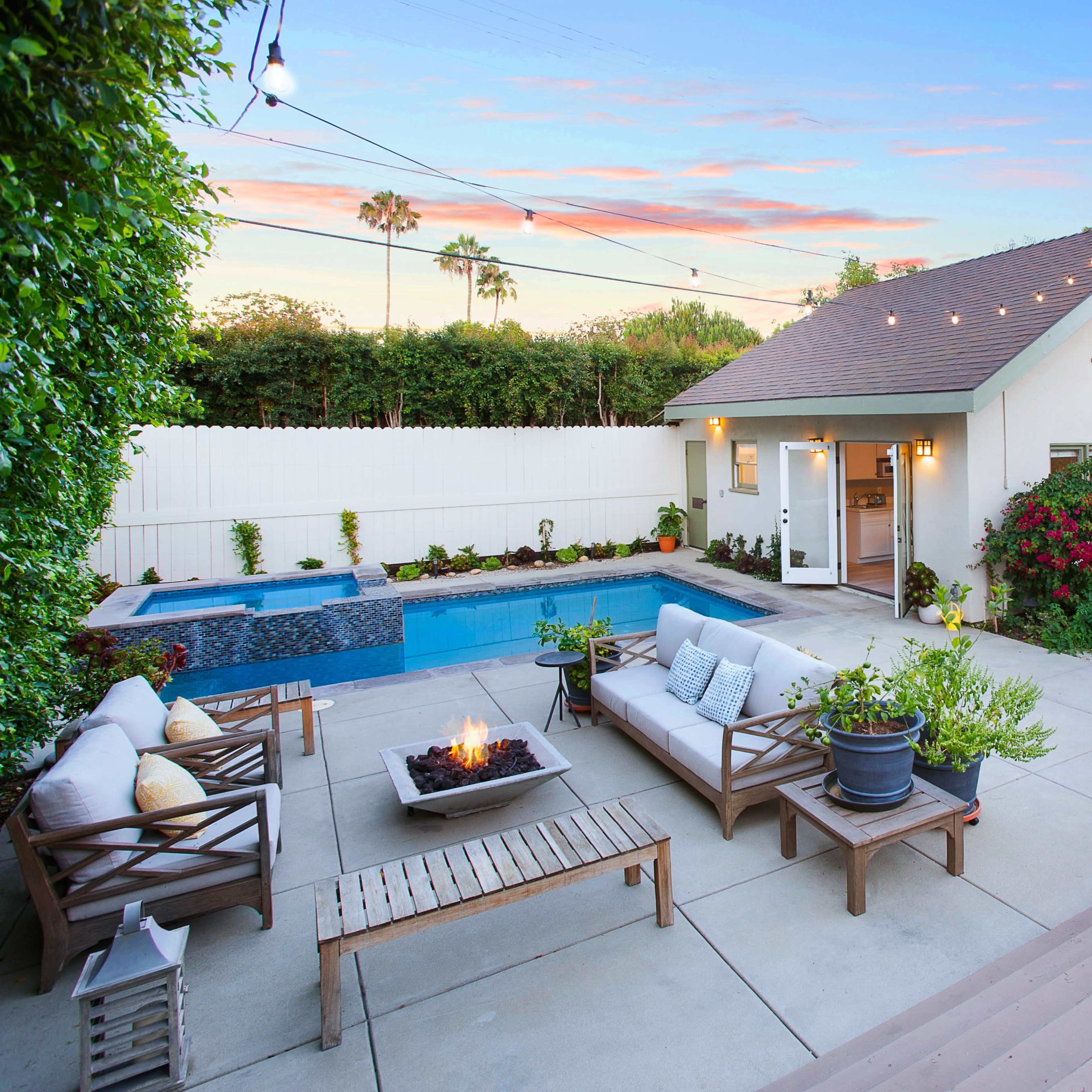 A backyard with a swimming pool, a seating area, and a small house surrounded by lush greenery and flowering plants at sunset.
