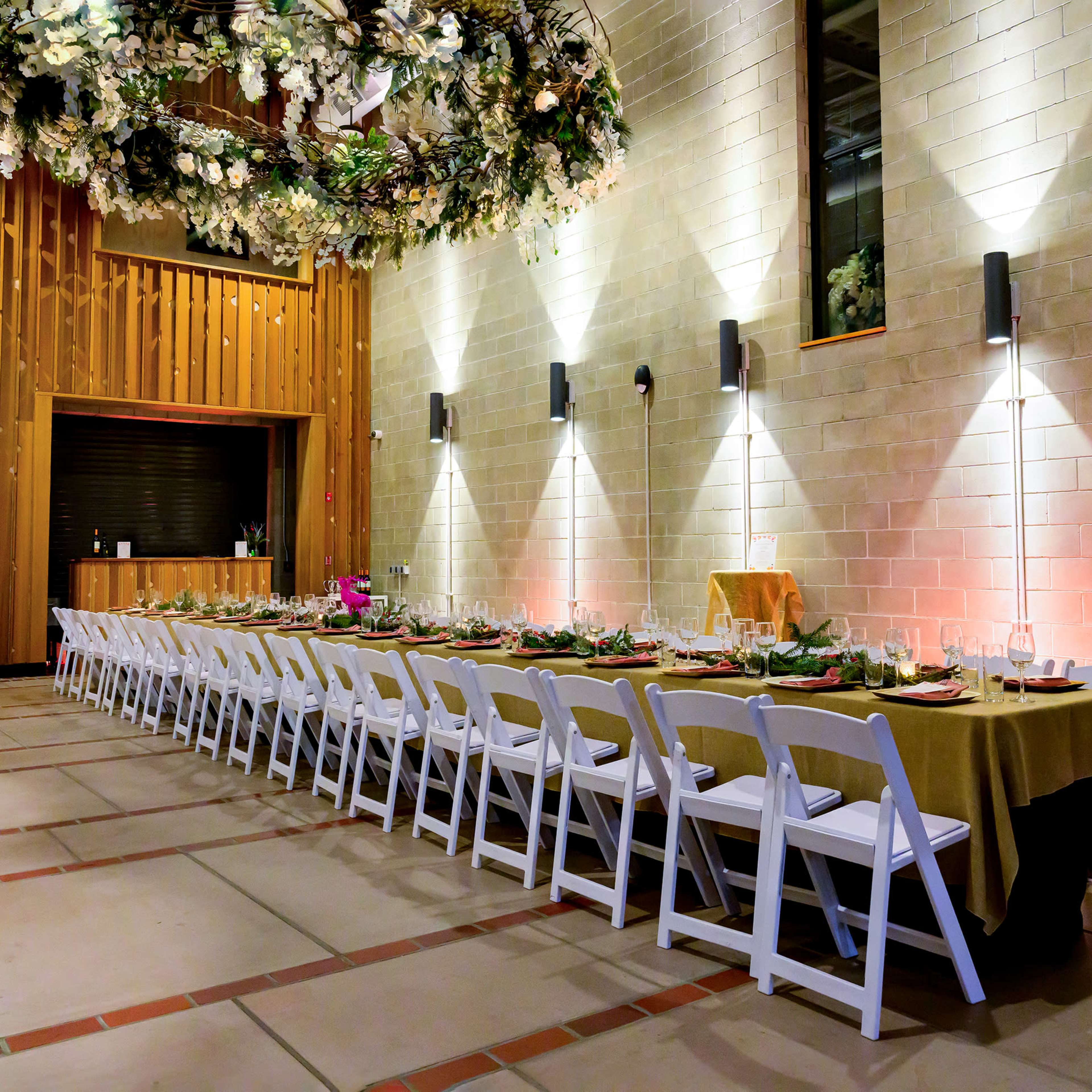 A long, elegantly set dining table in a spacious hall with wooden walls, decorated with floral arrangements and subtle lighting.