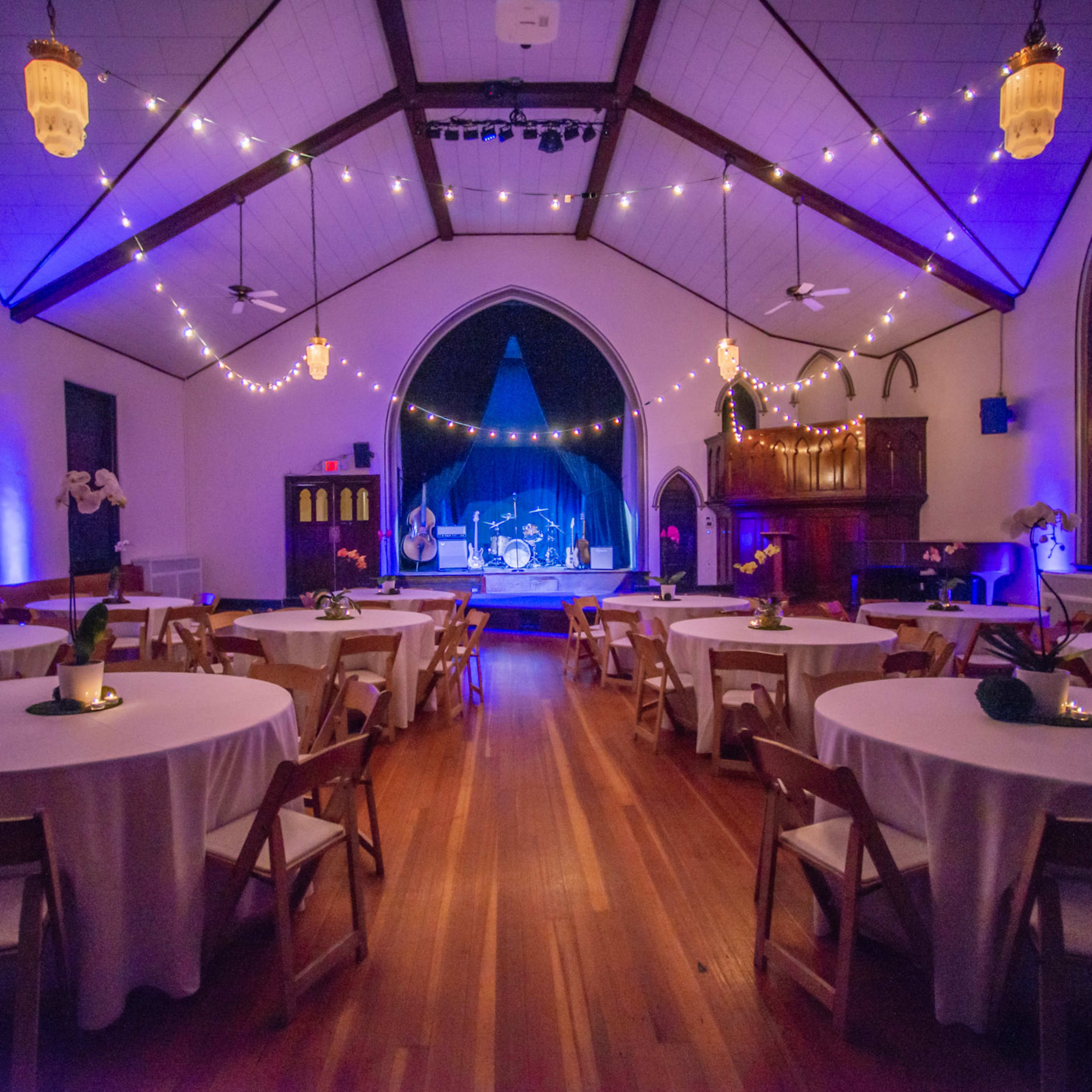 An elegantly decorated event space with round tables, wooden flooring, and a stage illuminated by soft blue and purple lighting.