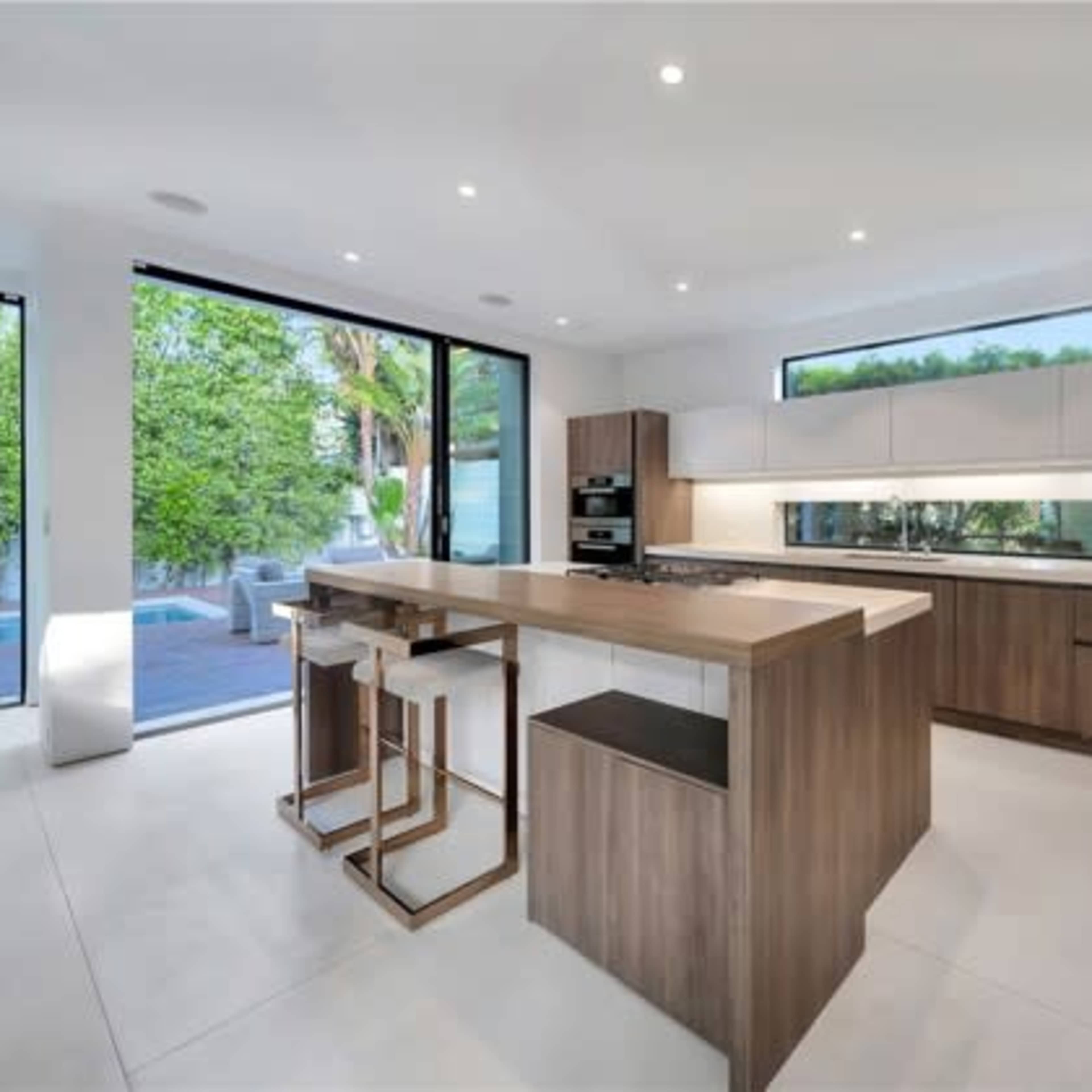 A modern kitchen features a large island with bar seating, sleek cabinetry, and large windows overlooking a patio and pool area.
