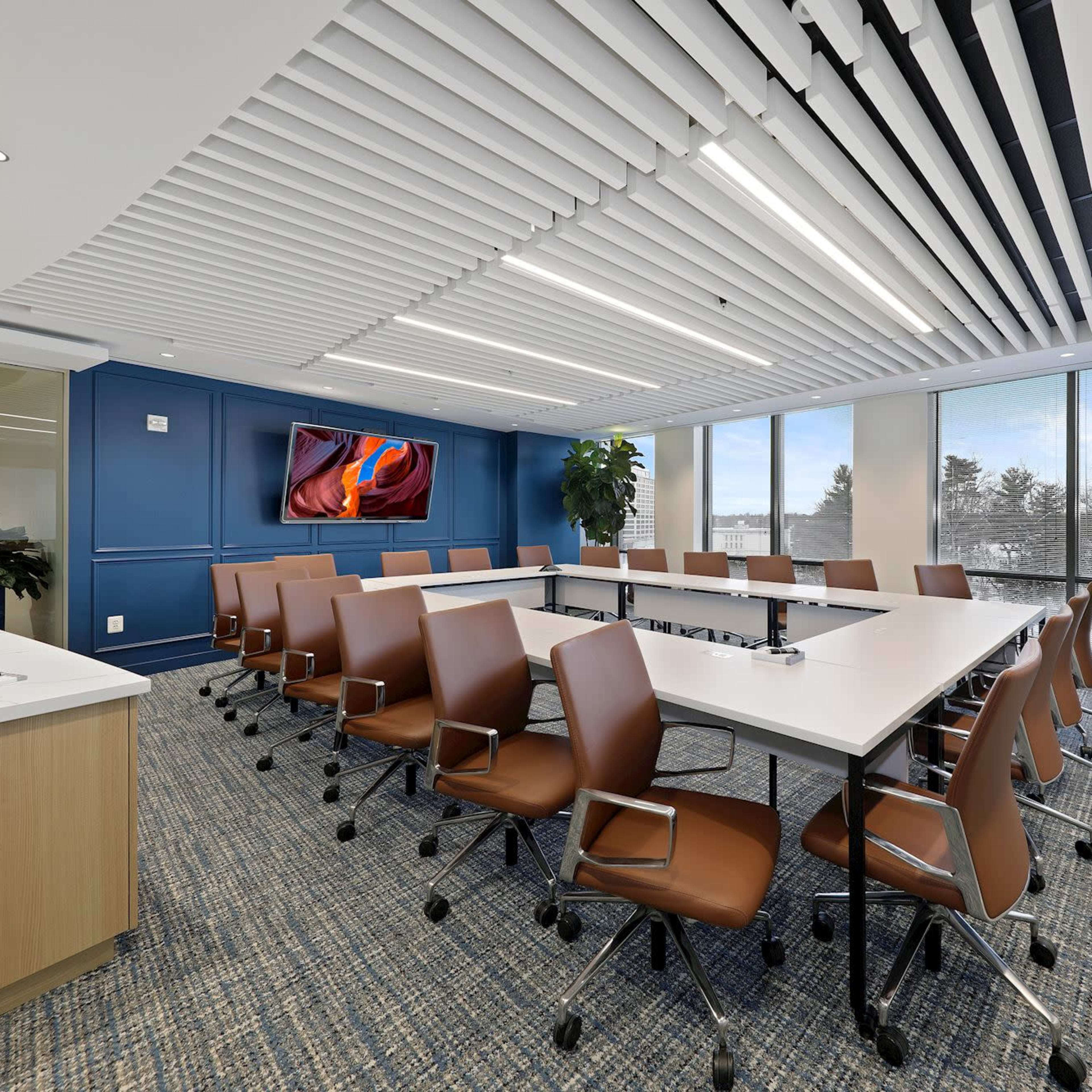A modern conference room features a long table surrounded by brown chairs, with large windows providing natural light and a television mounted on the wall.