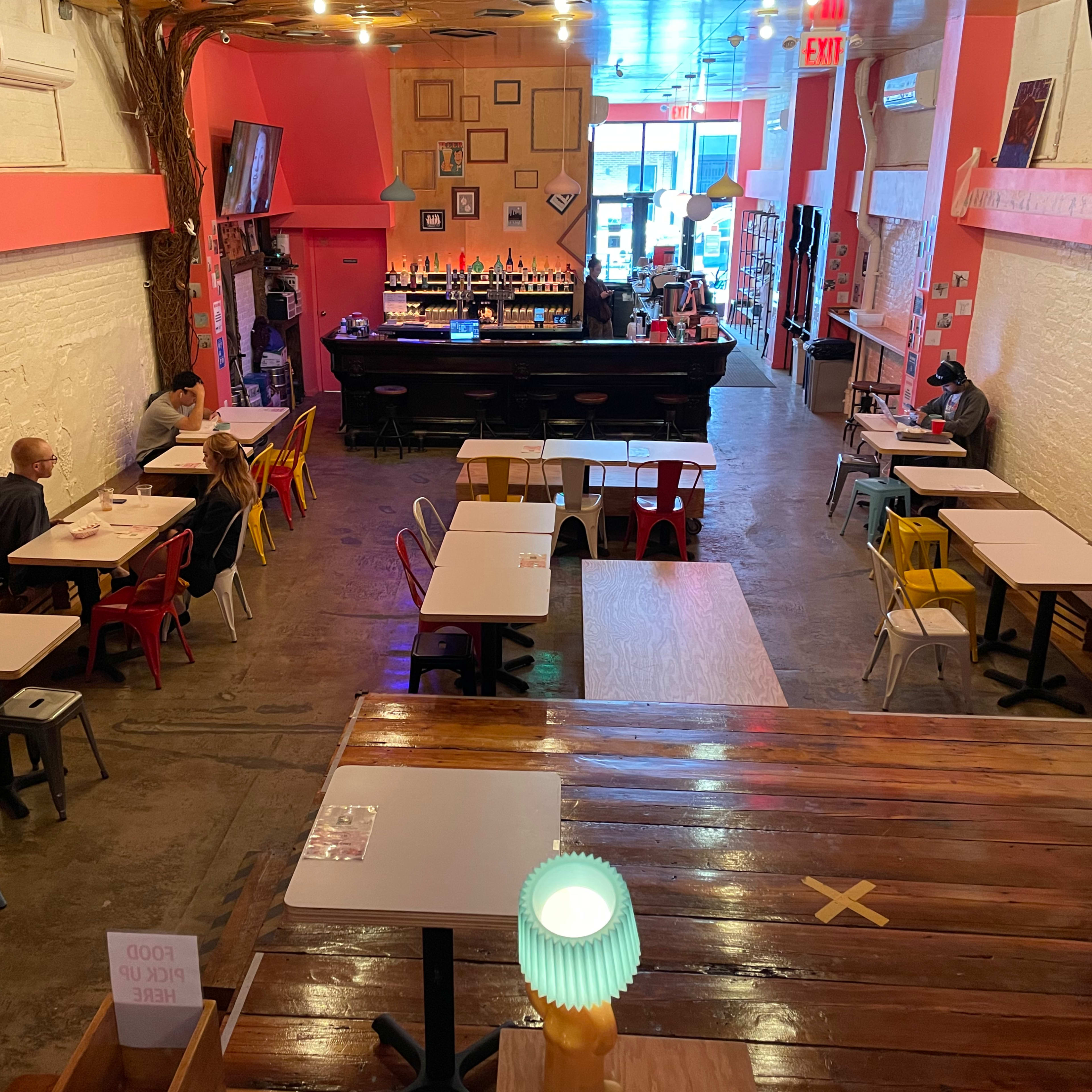 A brightly colored café interior with several small tables and chairs, a bar at the back, and patrons seated at the tables.