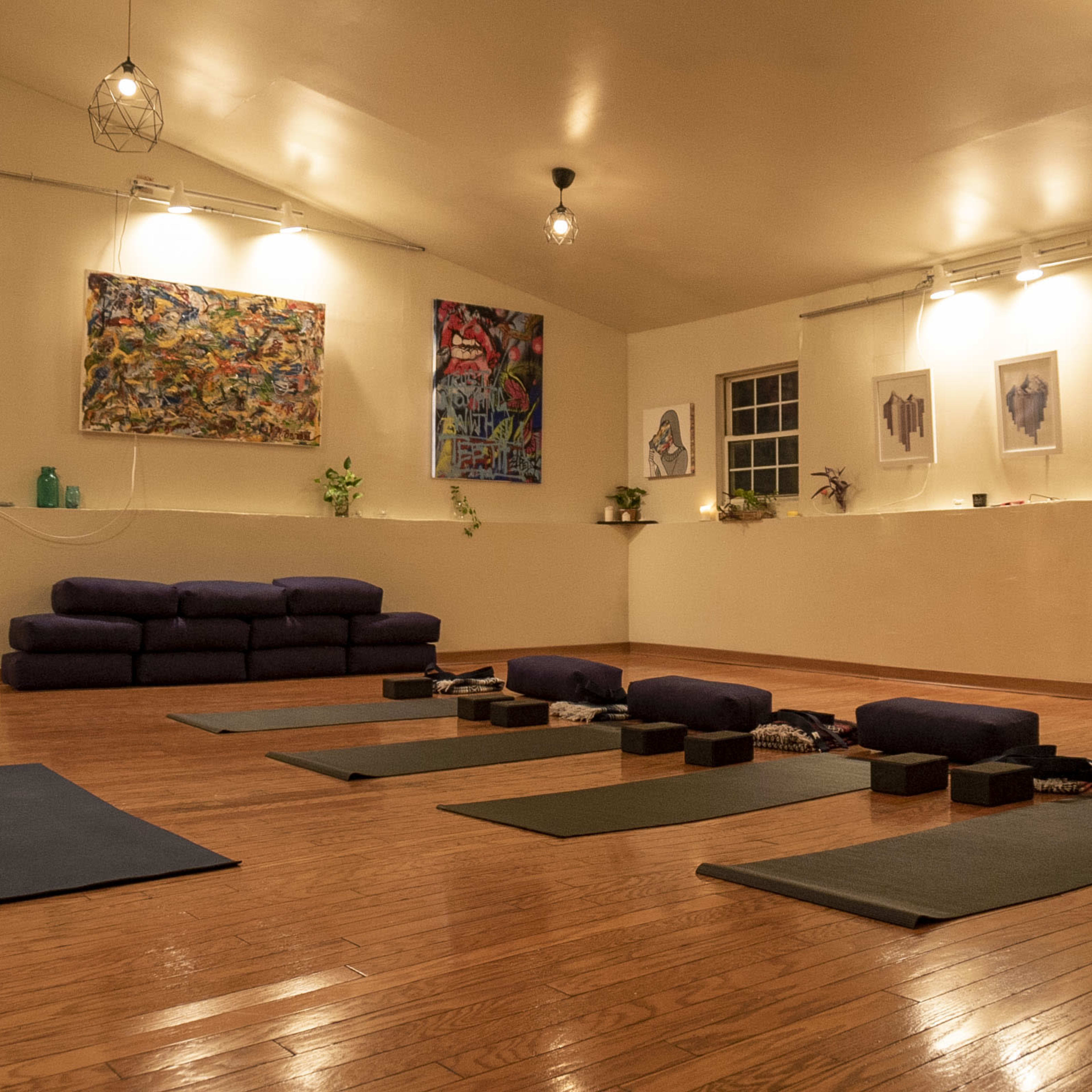 A spacious yoga studio with wooden floors, several yoga mats arranged in rows, and a couch along the back wall, adorned with artwork and plants.