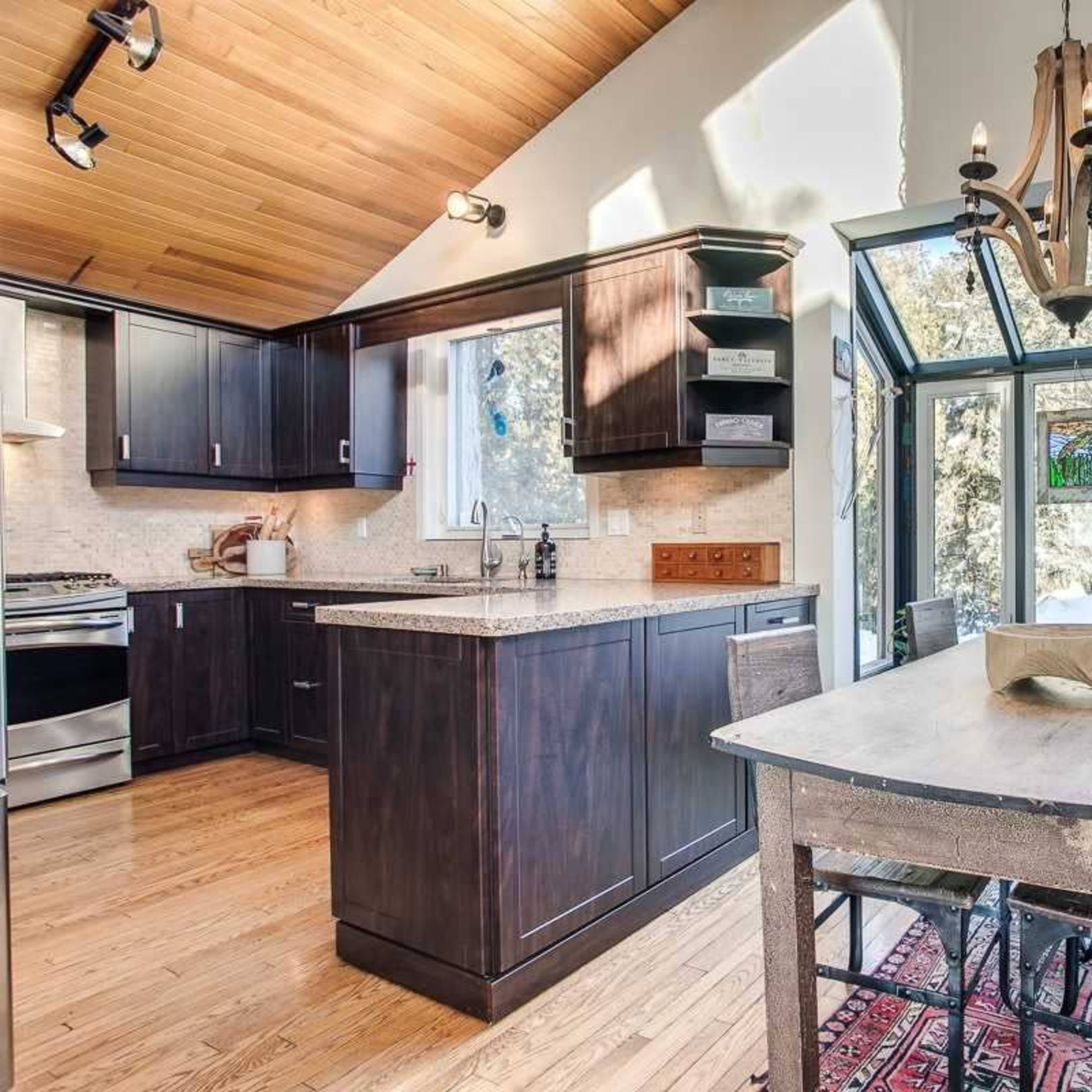 A modern kitchen featuring dark cabinetry, a granite countertop, and a dining area with a wooden table and chairs, illuminated by natural light from large windows.