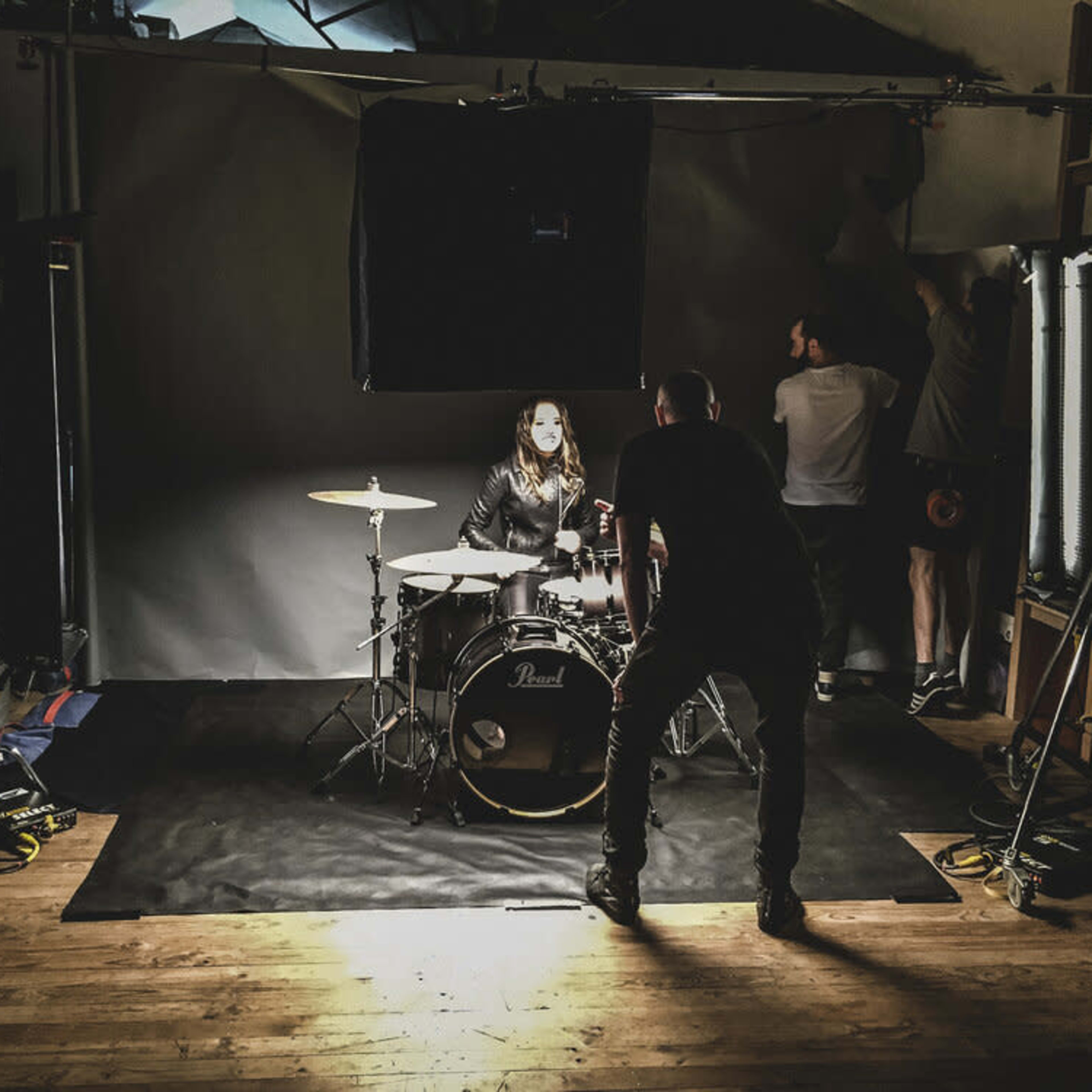 A drummer sits at a drum kit while a crew prepares the lighting and backdrop in a studio setting.