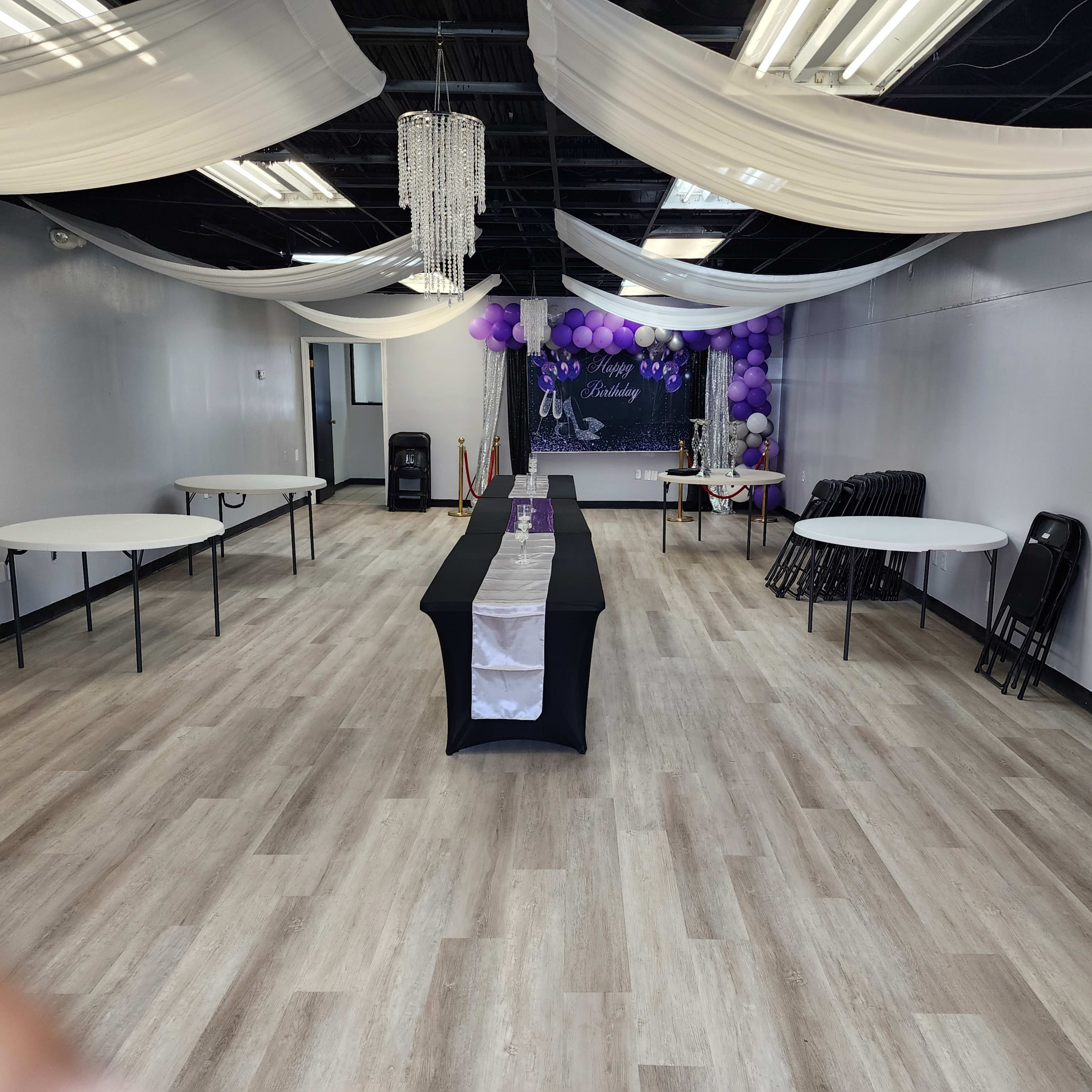 An empty event space with tables arranged around the room, adorned with purple balloons and draped fabric overhead.