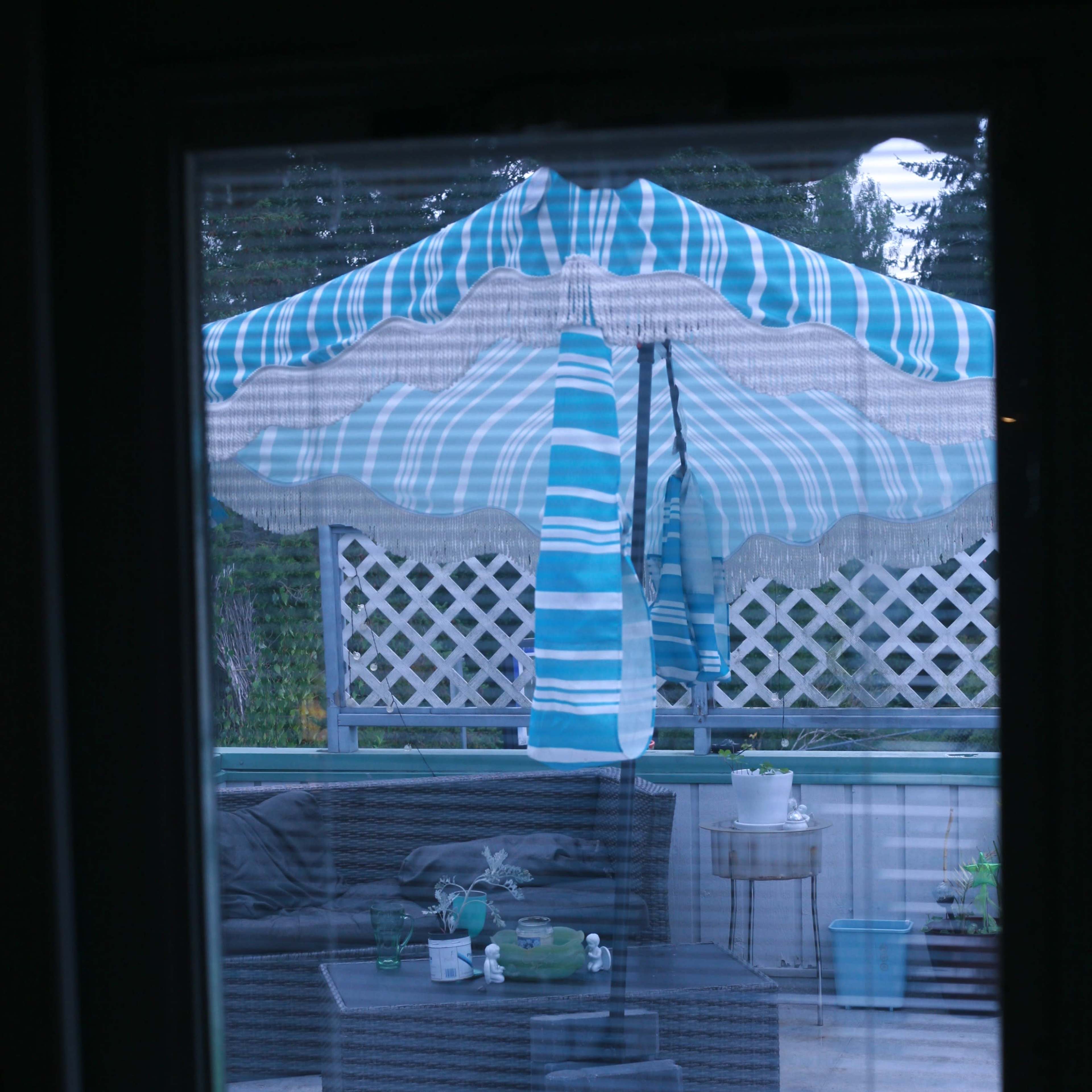 A striped umbrella stands over a patio table set with plants and drinkware, viewed through a window.