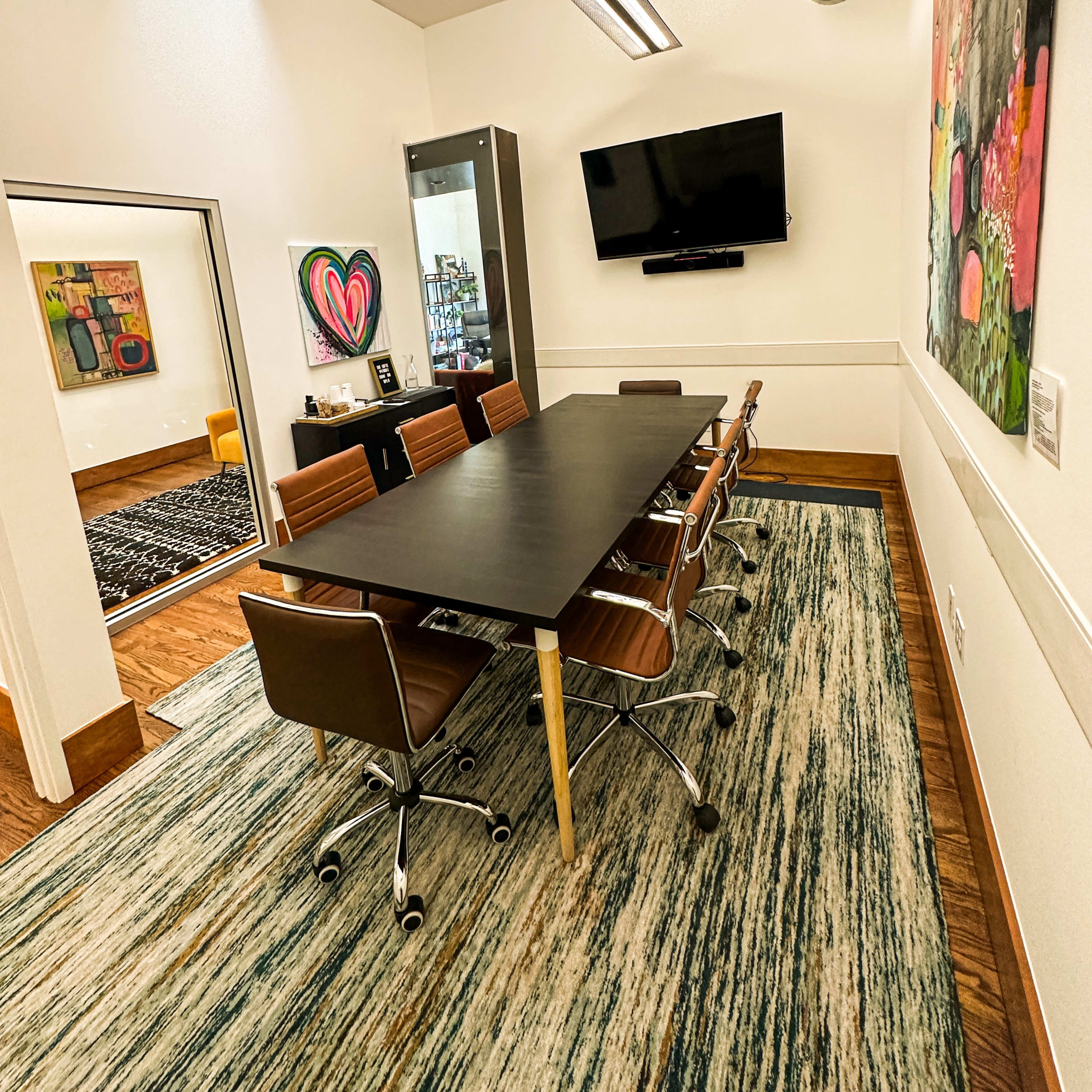 A modern conference room with a long table and six chairs, a wall-mounted TV, and colorful artworks on the walls.