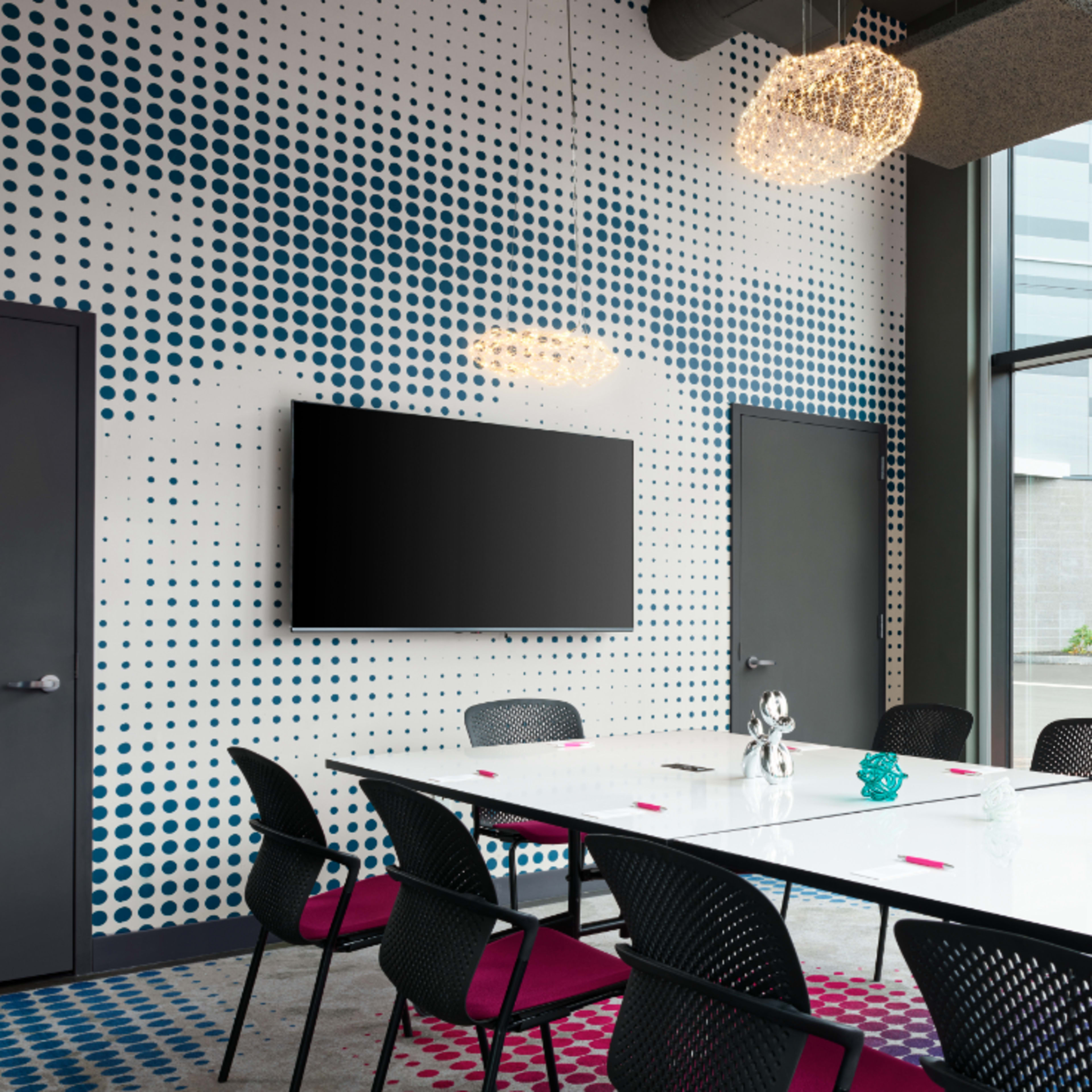 A modern conference room features a long white table surrounded by black chairs, with a large window displaying an outdoor view and a wall decorated with blue polka dots.