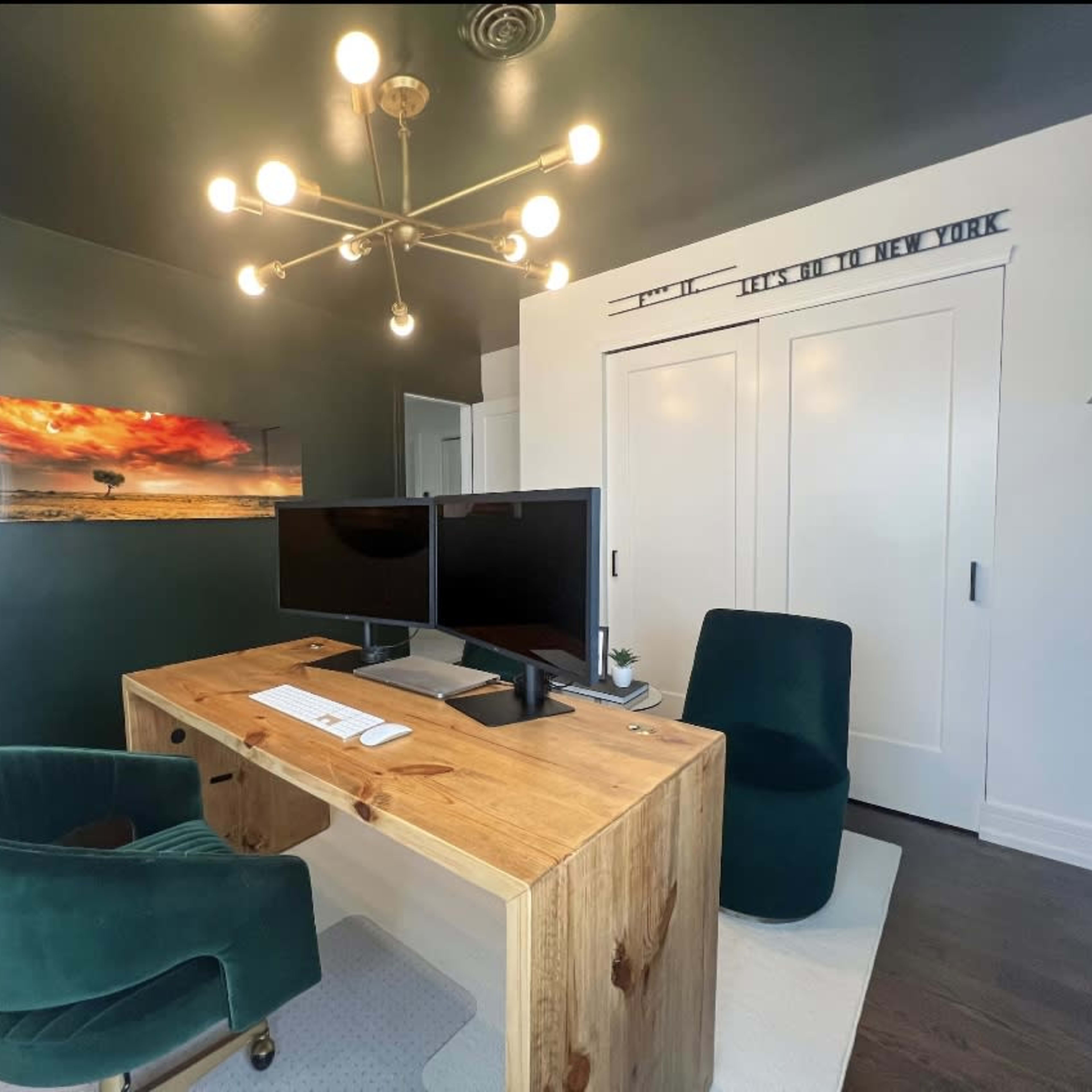 A modern office space features a wooden desk with two monitors, a green velvet chair, and a white carpet against a dark green wall, with a landscape painting and a closed closet in the background.