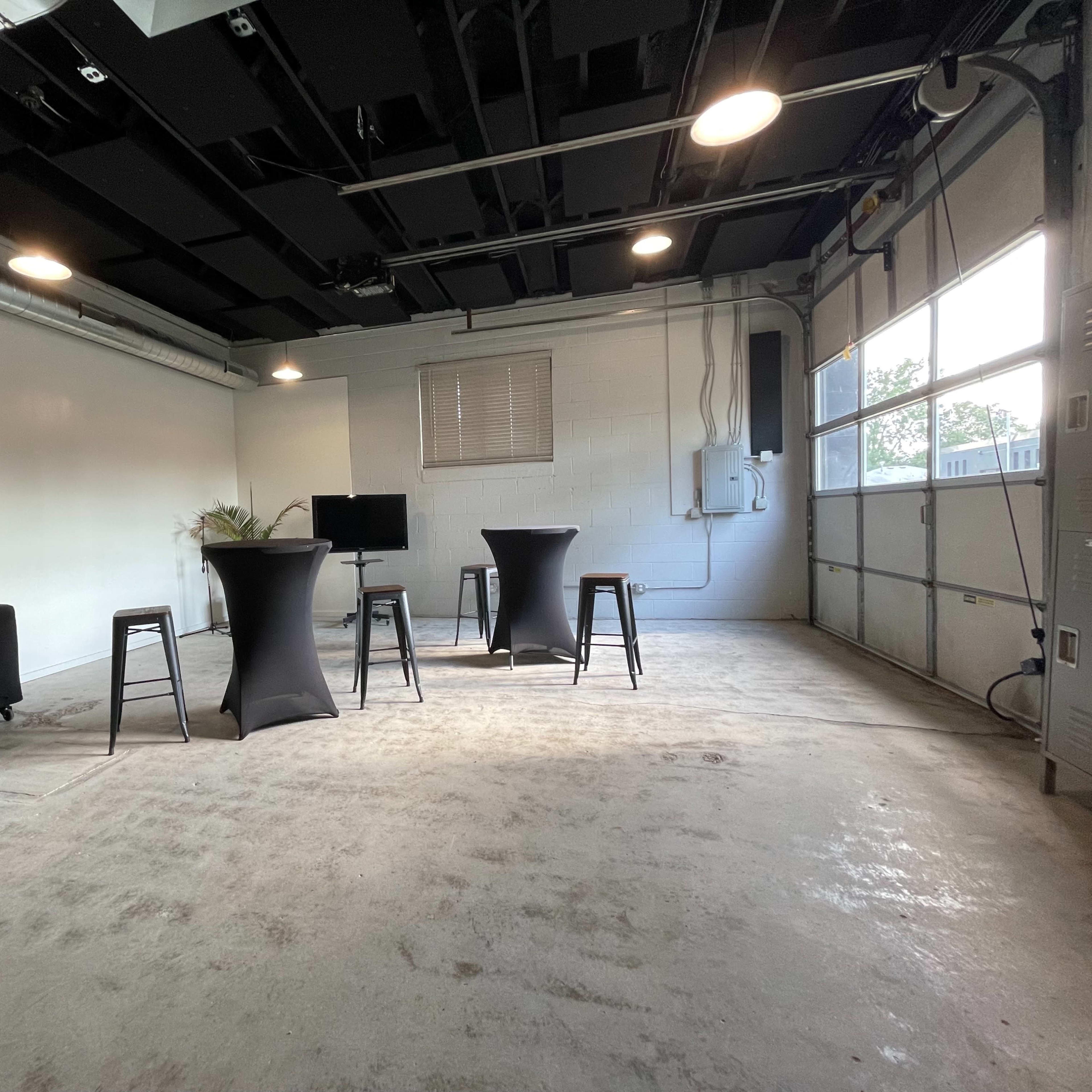 An empty garage with concrete flooring, featuring three high tables and a monitor on a stand, with a large window letting in natural light.
