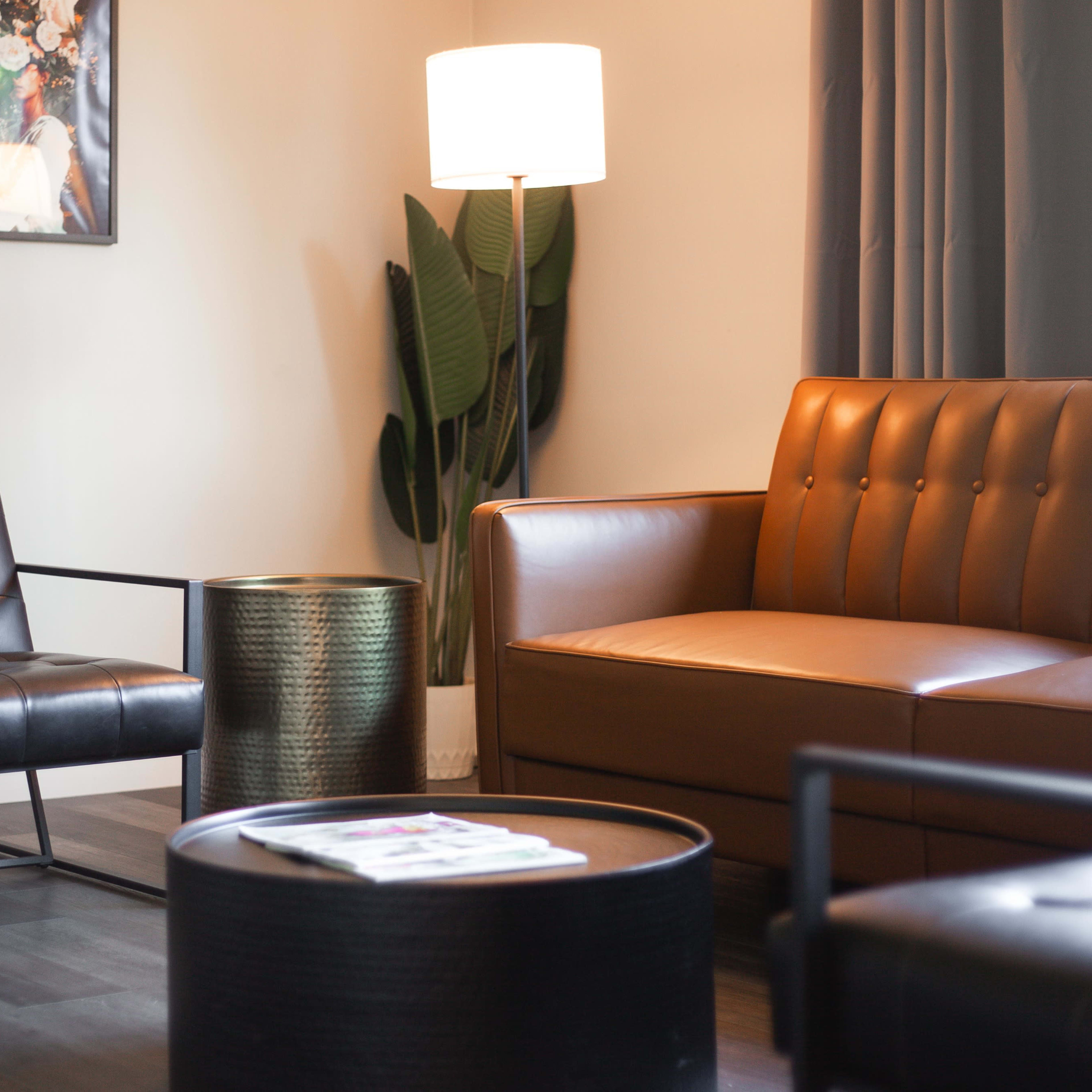 The scene features a modern living room with a leather sofa, a black accent chair, a round coffee table, and a standing lamp beside a potted plant.