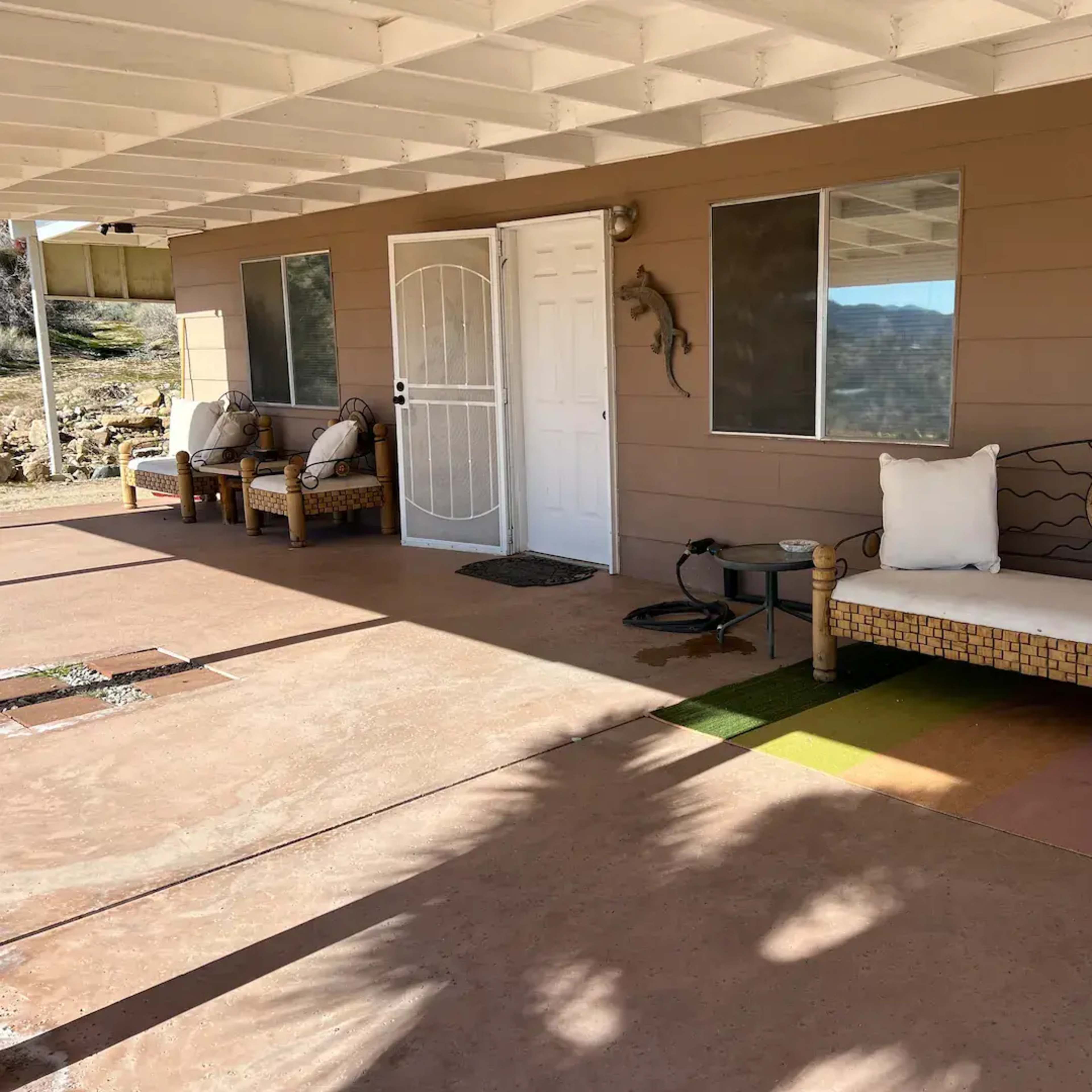 A covered porch features two wicker benches with cushions, a small table, and large windows leading into a tan-colored house.