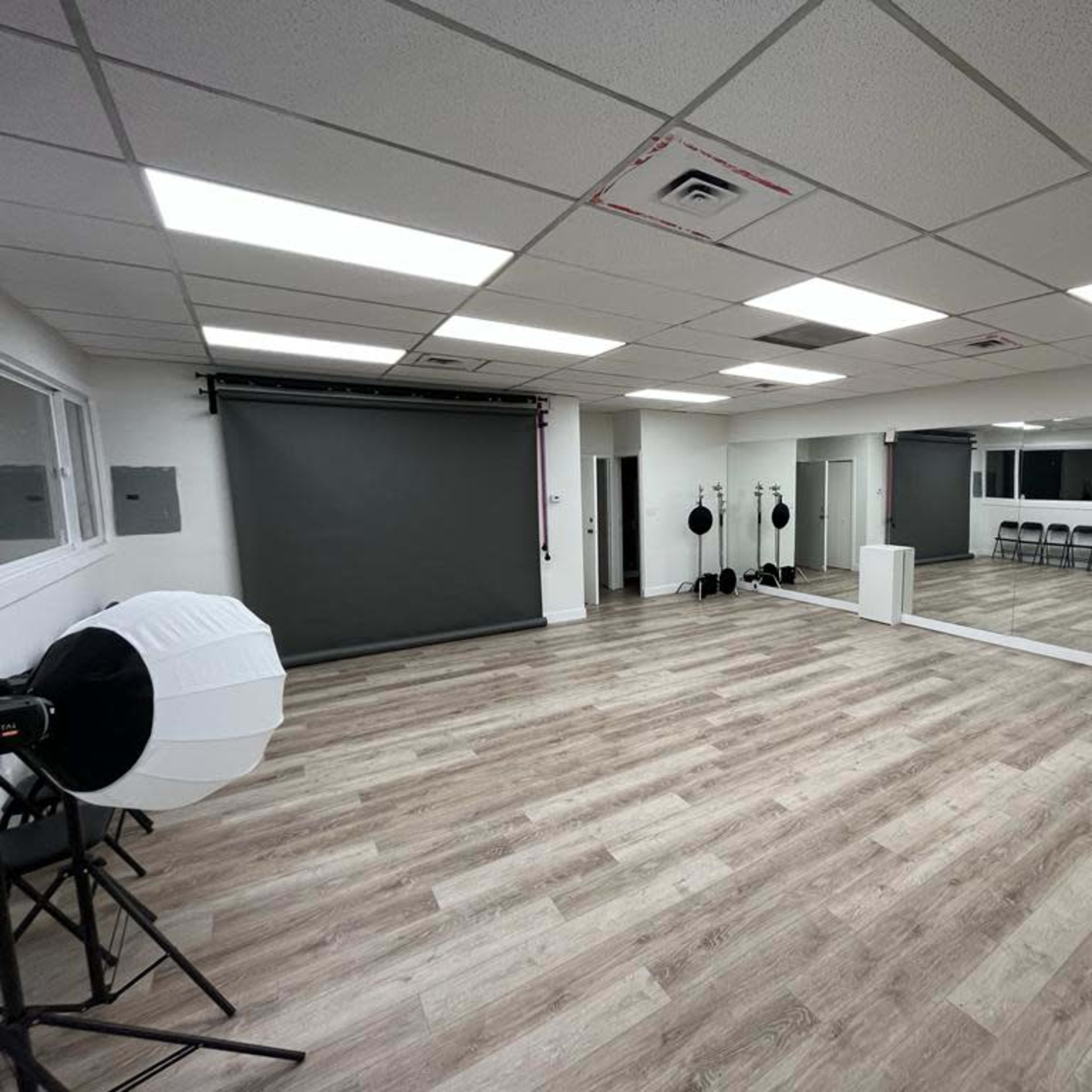 A spacious room with a mirrored wall, rolled-up backdrop, and photography lighting equipment arranged beside rows of folding chairs on a wooden floor.
